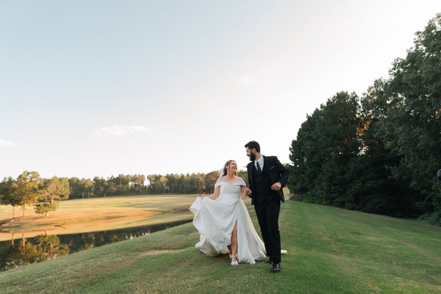 A couple walks hand in hand across a scenic hilltop meadow during golden hour at sunset.