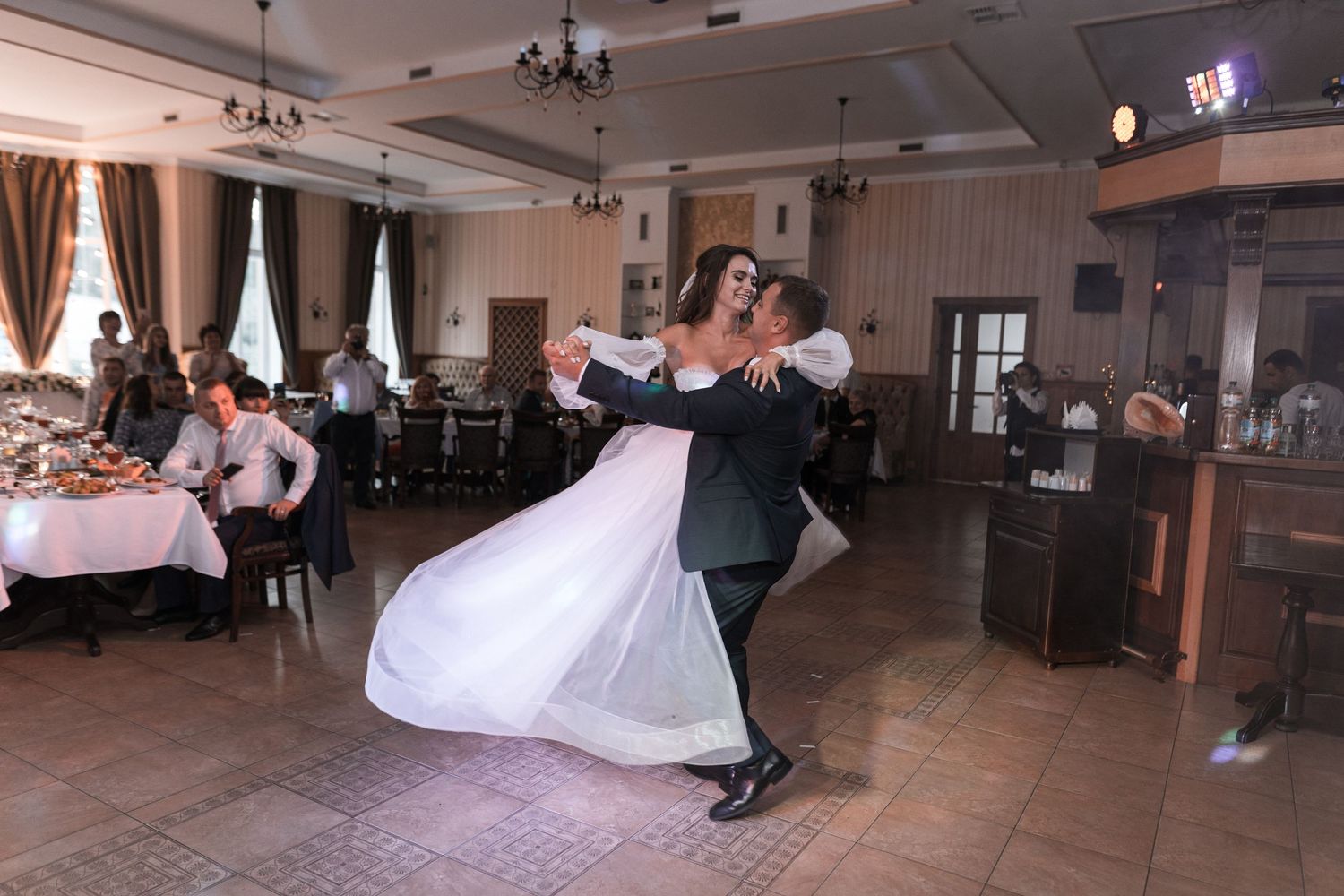 A couple performs a romantic dance dip in a wedding reception hall with pink table settings and chandeliers.
