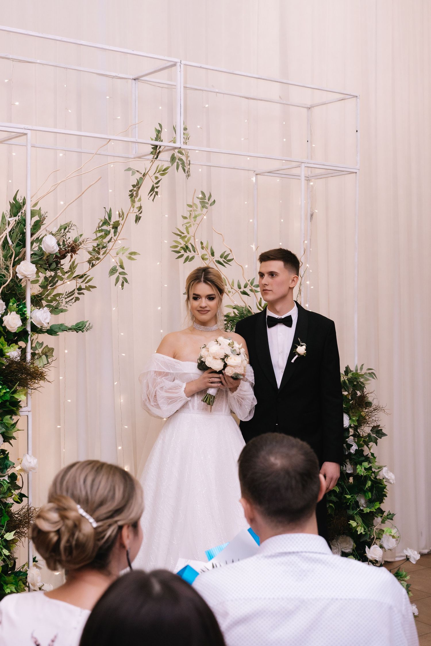 A couple in wedding attire perform their first dance in front of guests at an indoor reception venue with floral decor.