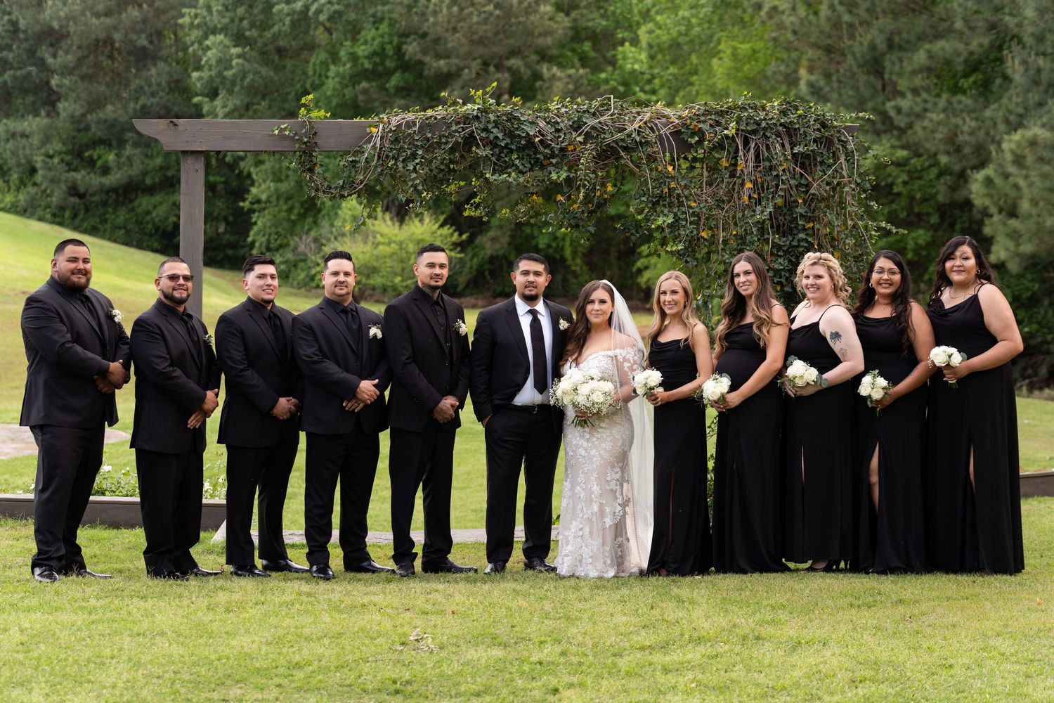 Wedding party poses creatively in line formation on grassy lawn for outdoor group photo shoot.