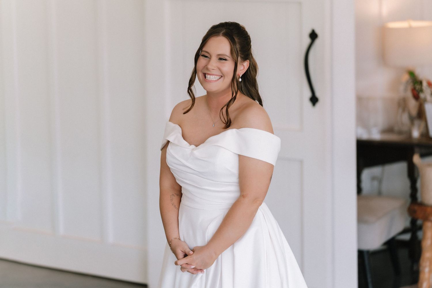 A joyful bride in an off-shoulder white wedding dress smiles while standing against a light-colored wall.