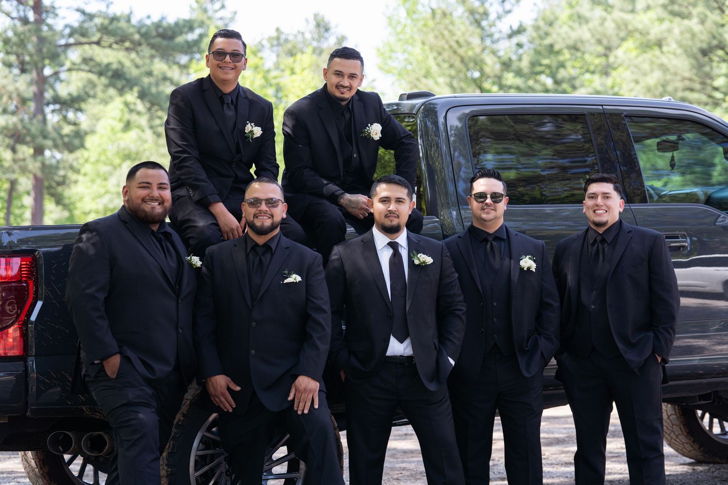 A group of groomsmen in black suits and sunglasses pose together in front of a black pickup truck outdoors.