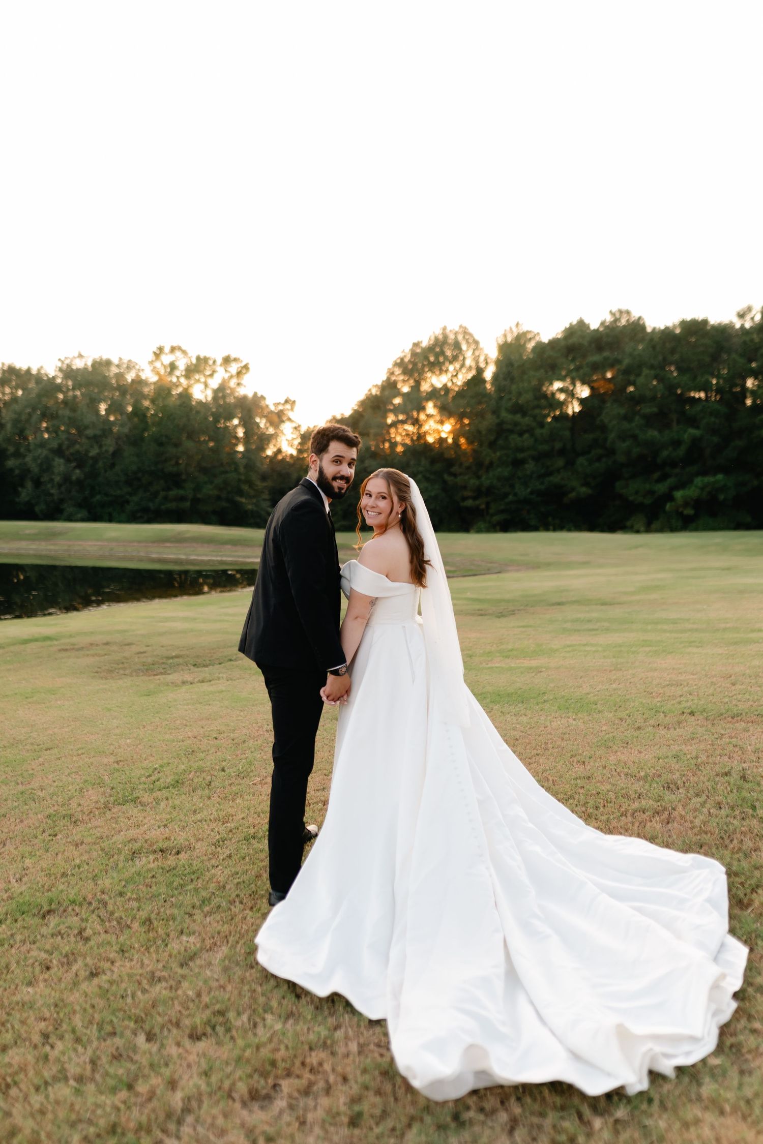 Bride and groom holding hands at sunset on a green lawn, bride wearing an elegant off-shoulder gown with long train.