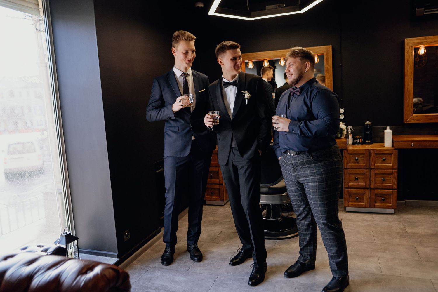 Wedding party members in navy suits gather near a window in a getting-ready room.