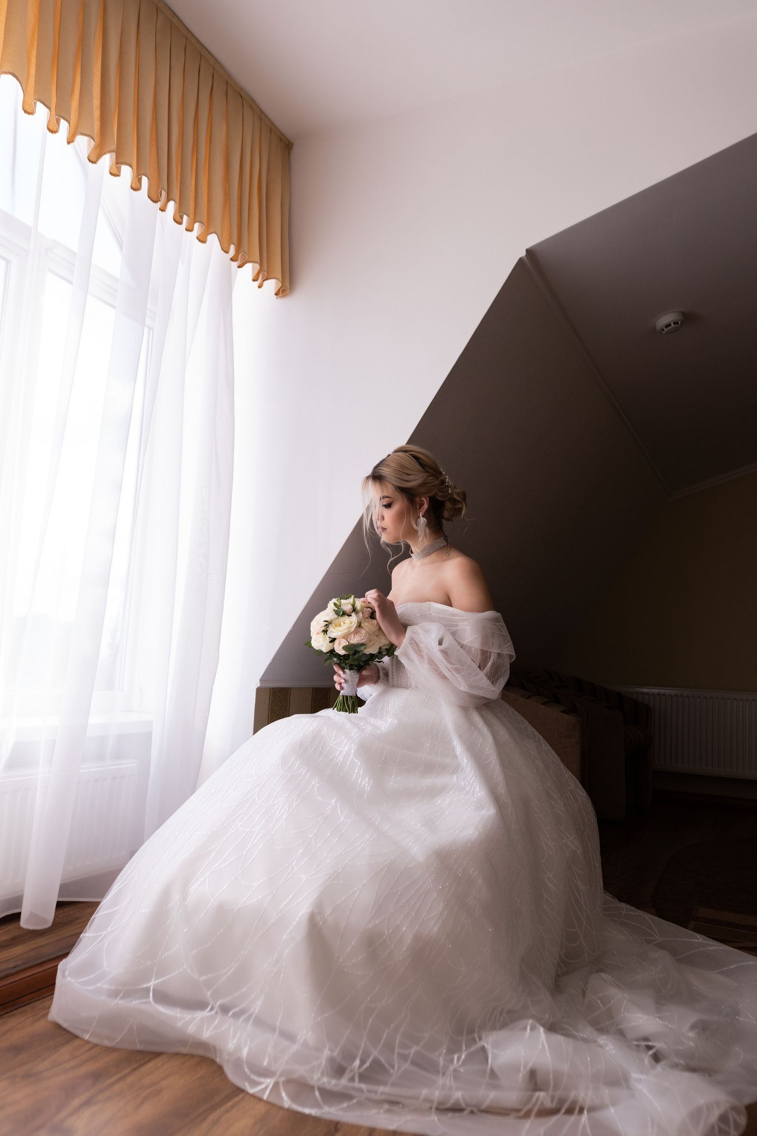 A bride in an elegant white off-shoulder ball gown sits gracefully by a window in soft natural light.