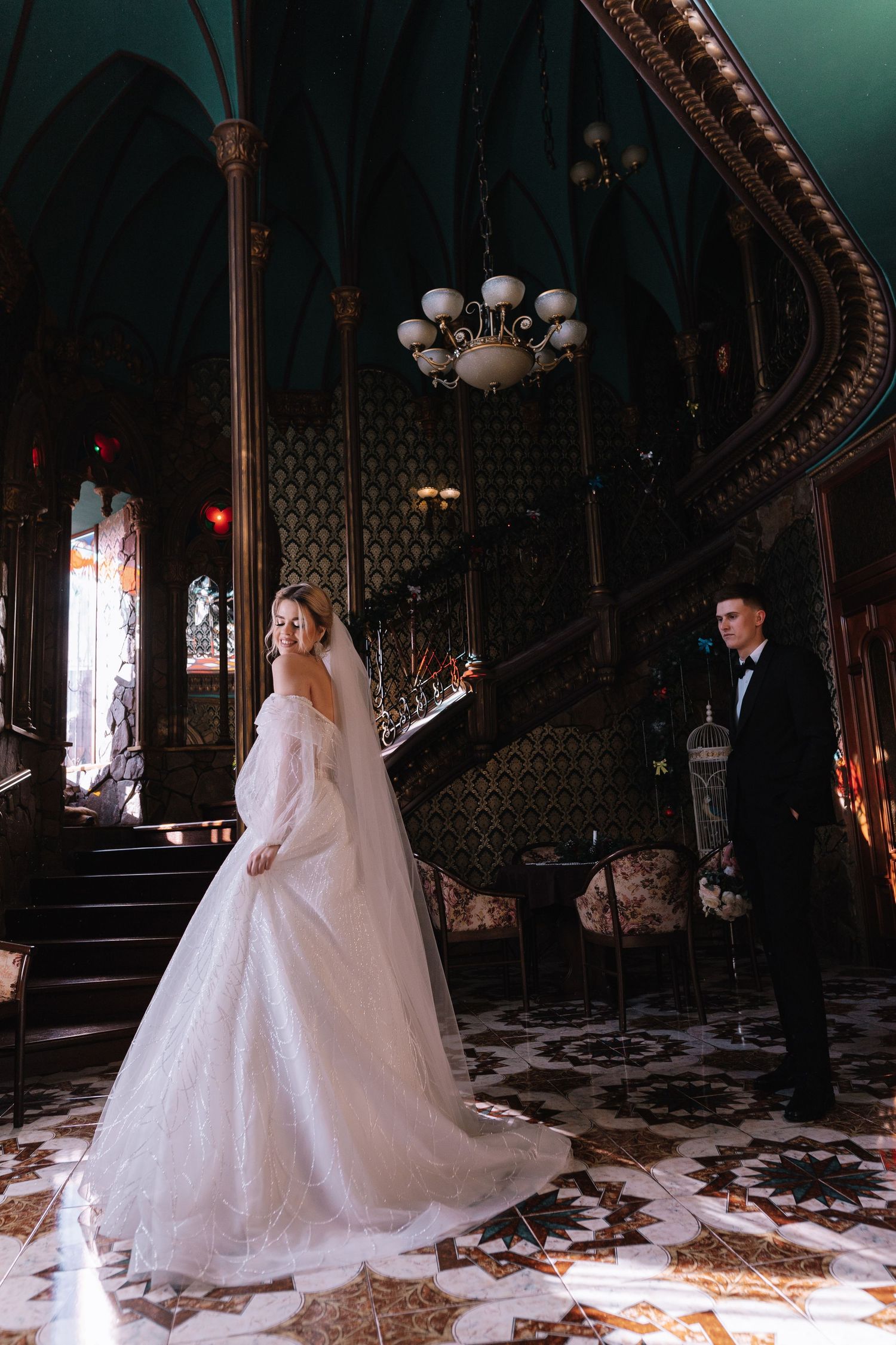 Elegant wedding photo in a vintage mansion interior with ornate staircase, chandelier and dramatic lighting.