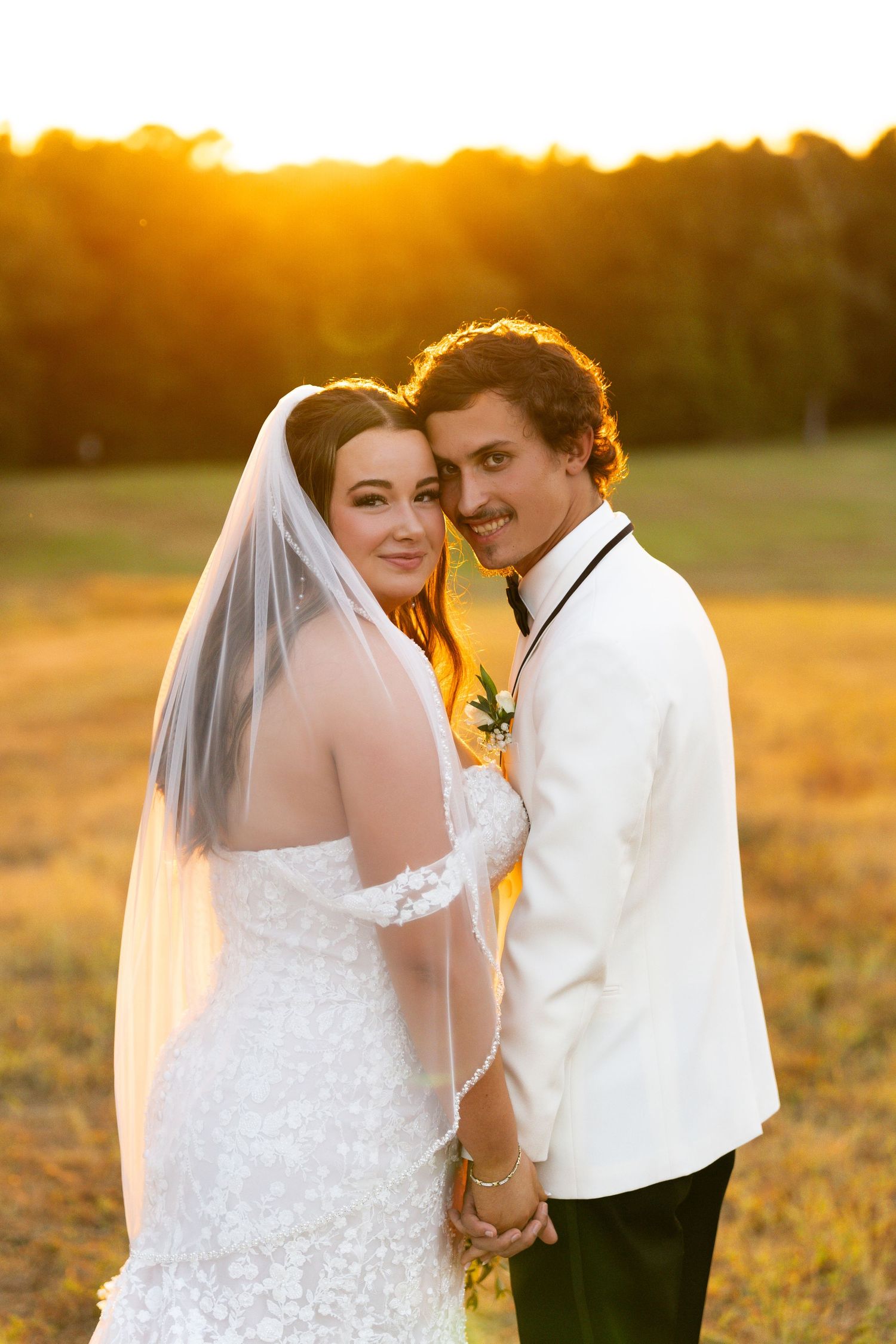 Romantic wedding couple embraces in golden sunlit field during sunset while wearing white formal attire and veil.
