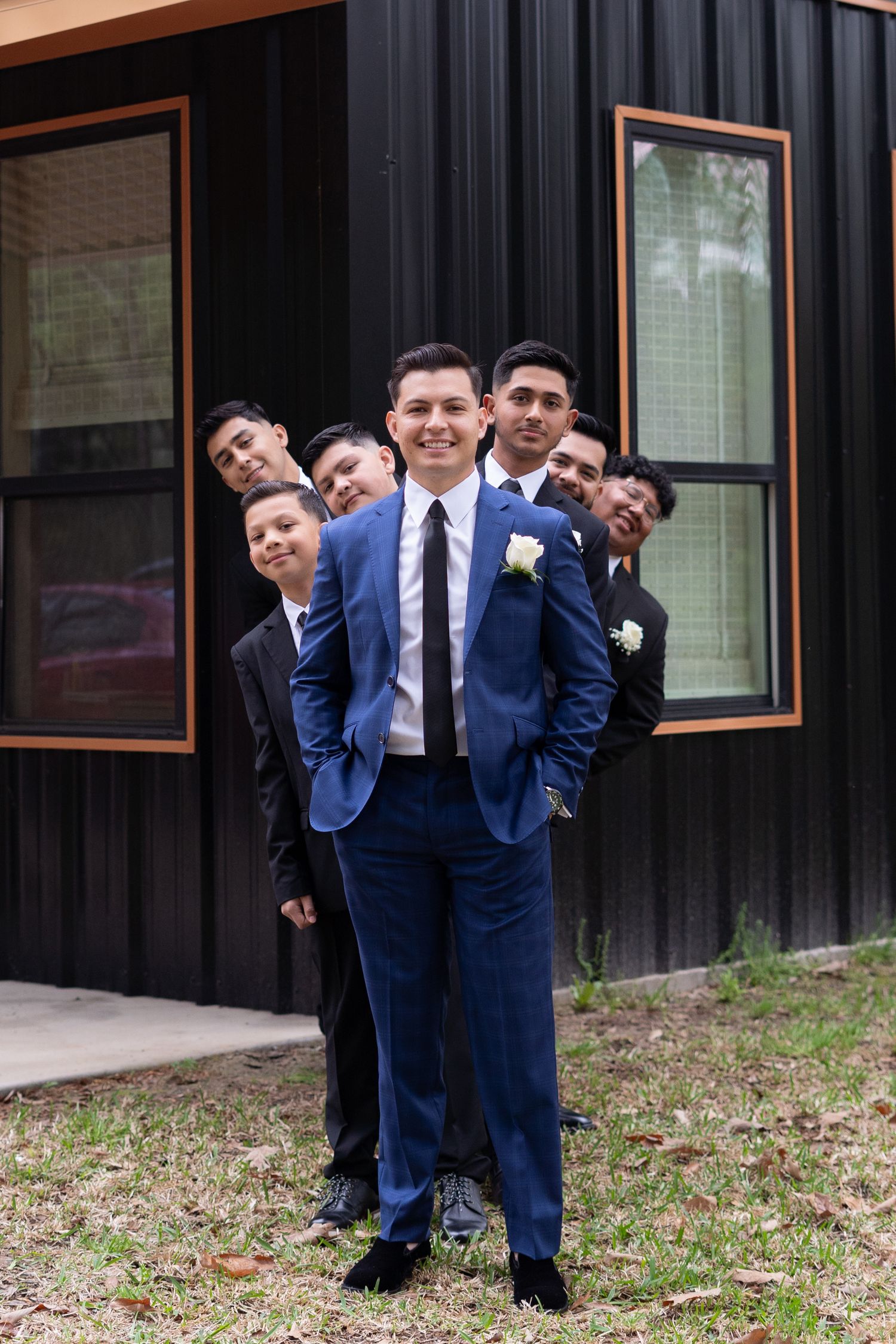 A group of groomsmen in suits pose playfully in a line behind the groom in a bright blue suit outside a building.