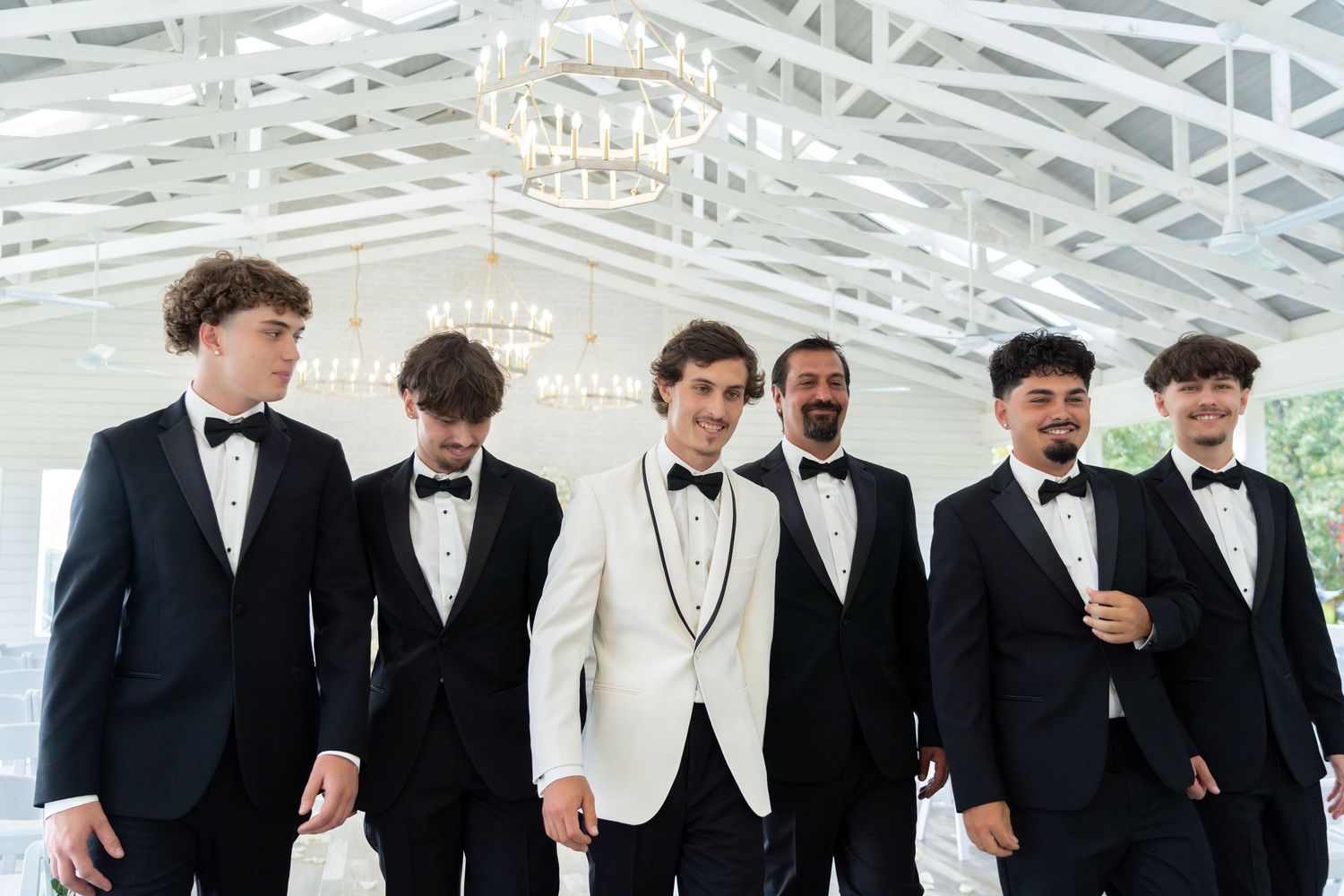 A group of groomsmen in black tuxedos and bow ties stand together smiling at a formal wedding event.