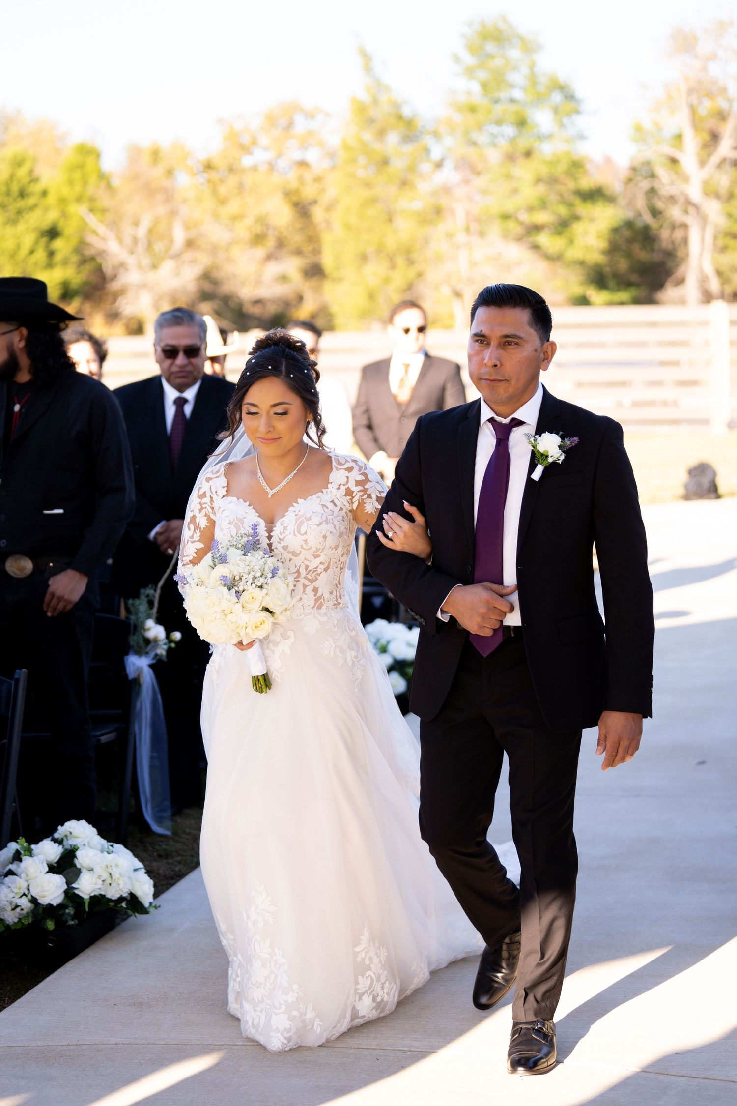 A bride in a white lace gown walks down an outdoor pathway with two people escorting her during a wedding ceremony.