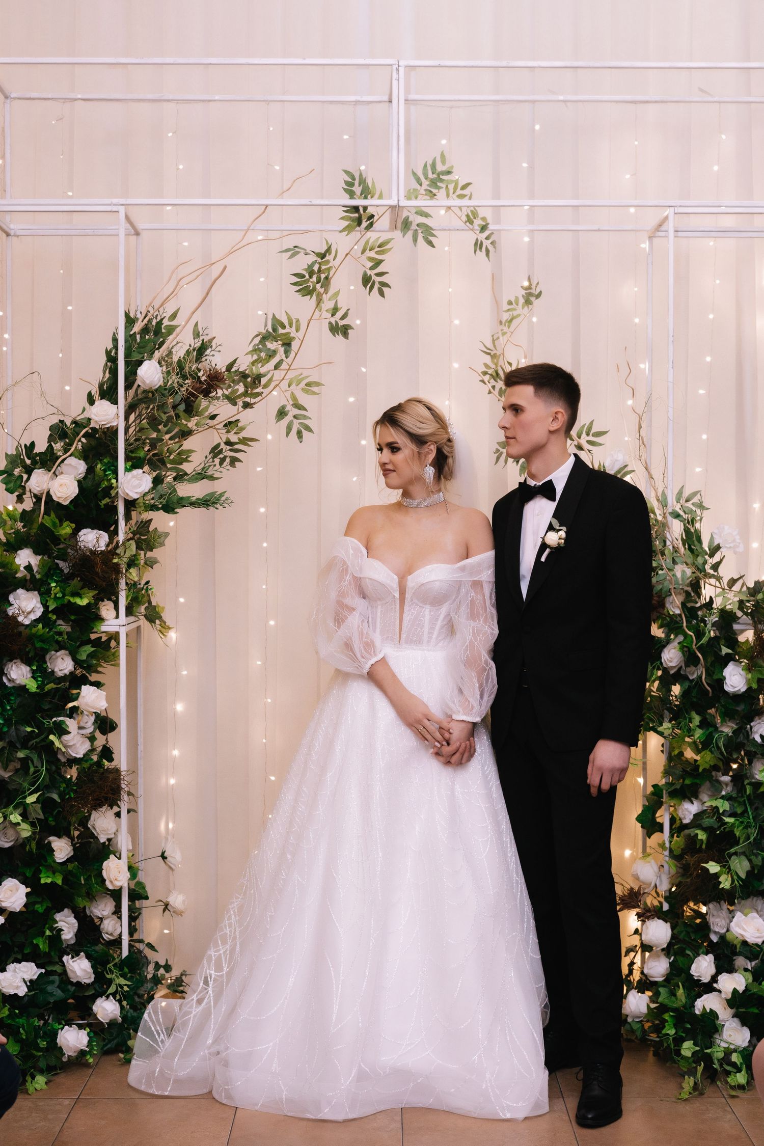 A romantic wedding couple poses against a blush backdrop adorned with elegant white floral arrangements and twinkling lights.