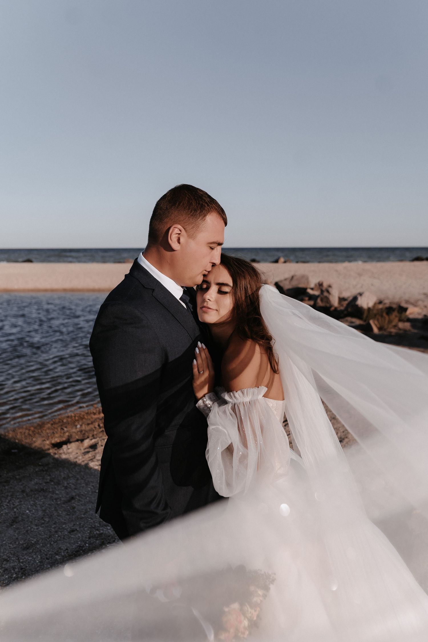 A romantic wedding couple embraces by the ocean at sunset as the bride's veil flows in the breeze.