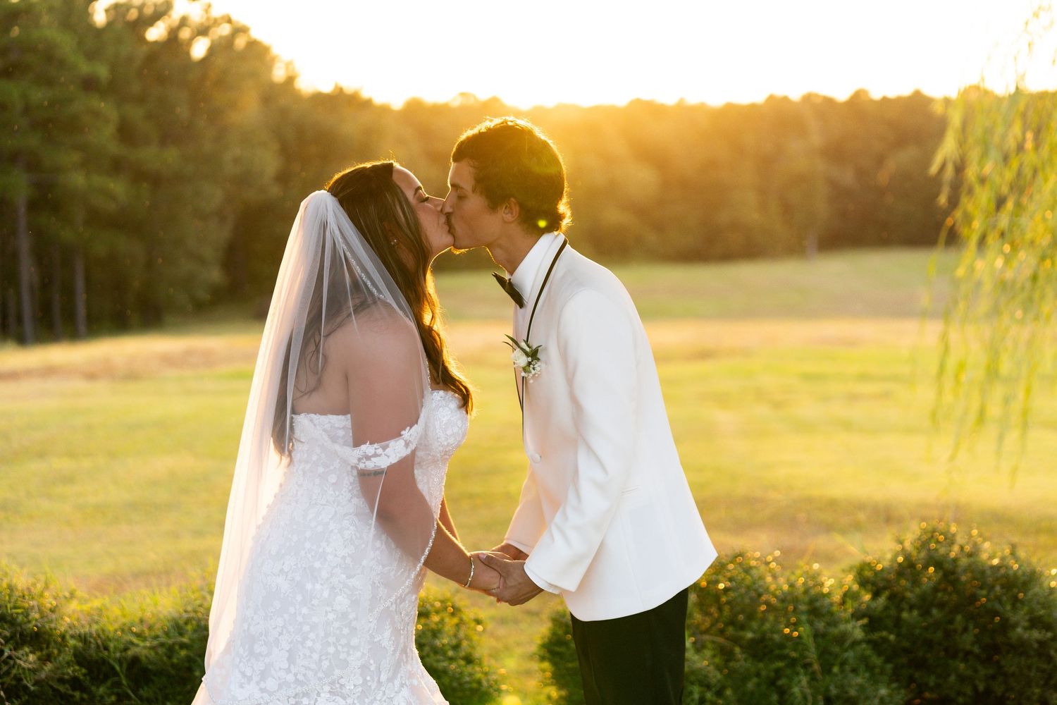 Romantic couple embraces during golden hour in a scenic outdoor field with sunlight glowing through trees.