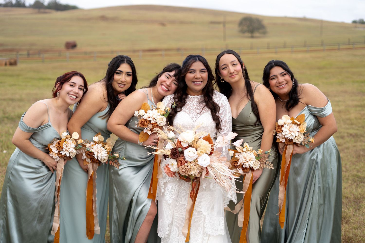 Bridesmaids in sage green dresses pose with bride holding autumn-colored bouquets in an outdoor field.