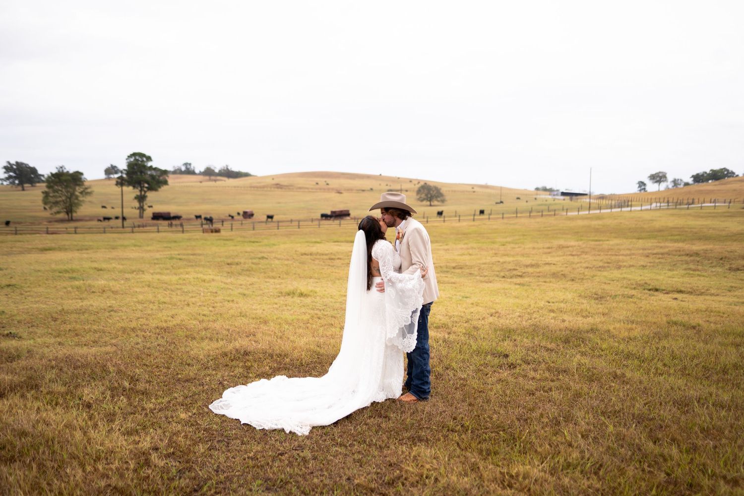 A couple in wedding attire share a romantic moment in a rustic field with rolling hills in the background.