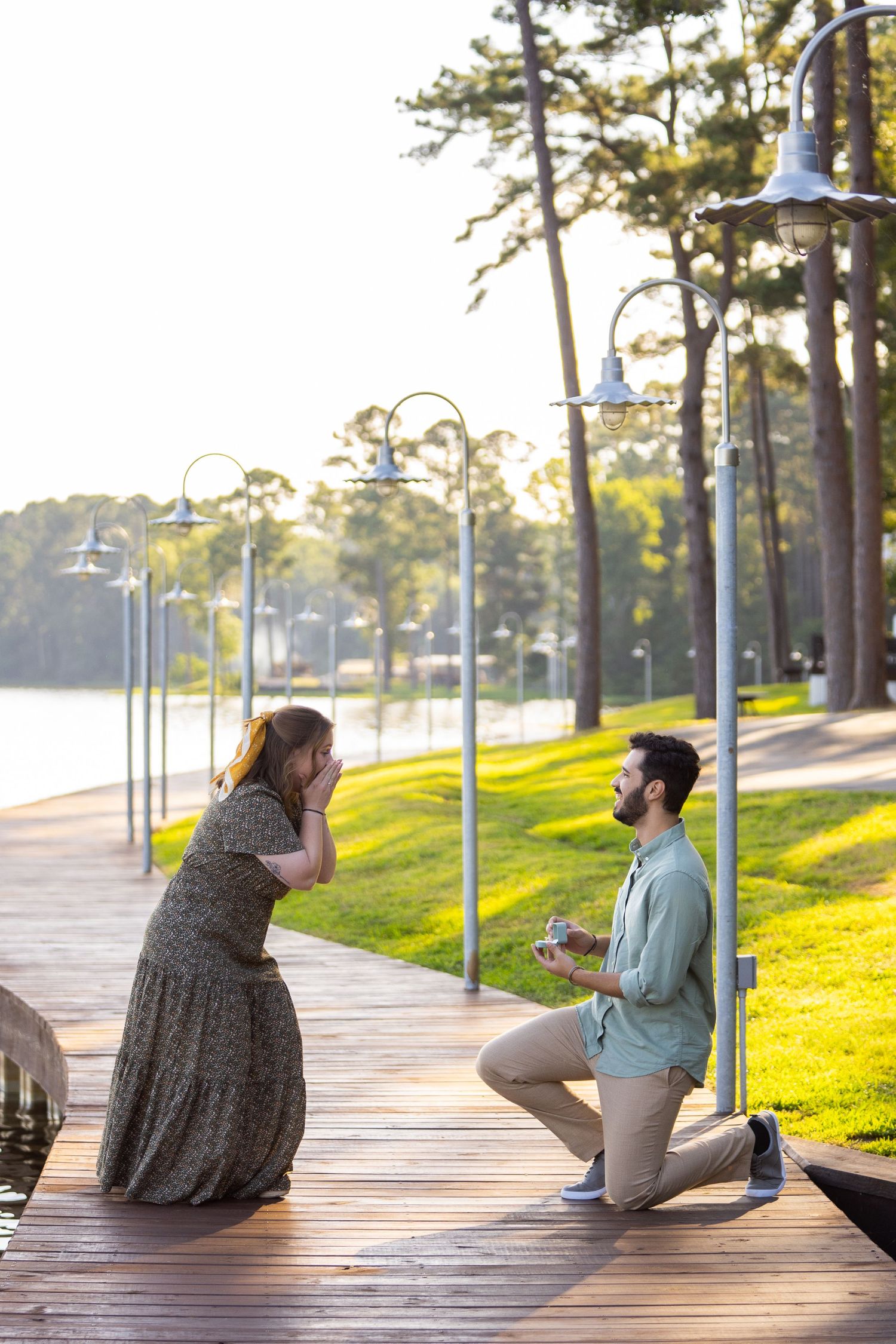 A romantic proposal moment on a wooden boardwalk with decorative lamp posts and pine trees at sunset.