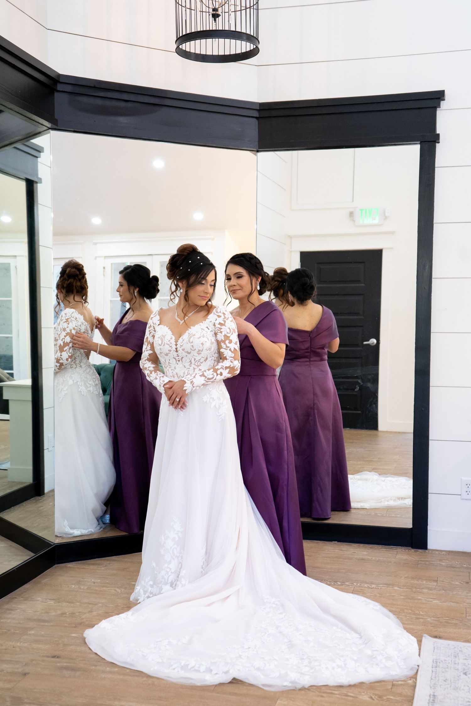 Bridesmaids in purple dresses help adjust the bride's long white lace train in a bright indoor space.
