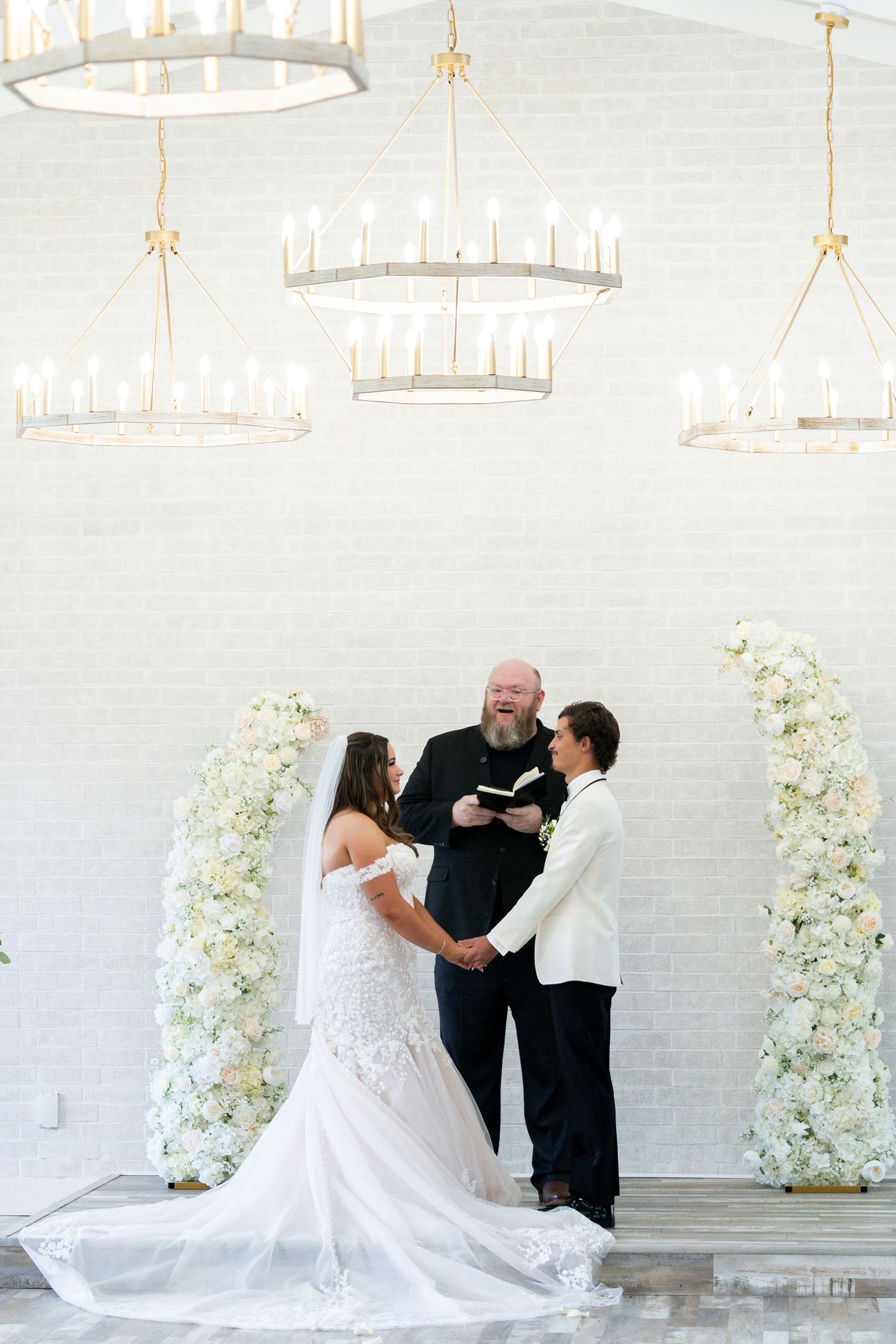 An elegant wedding ceremony setup with white floral arches and crystal chandeliers in a modern white interior space.