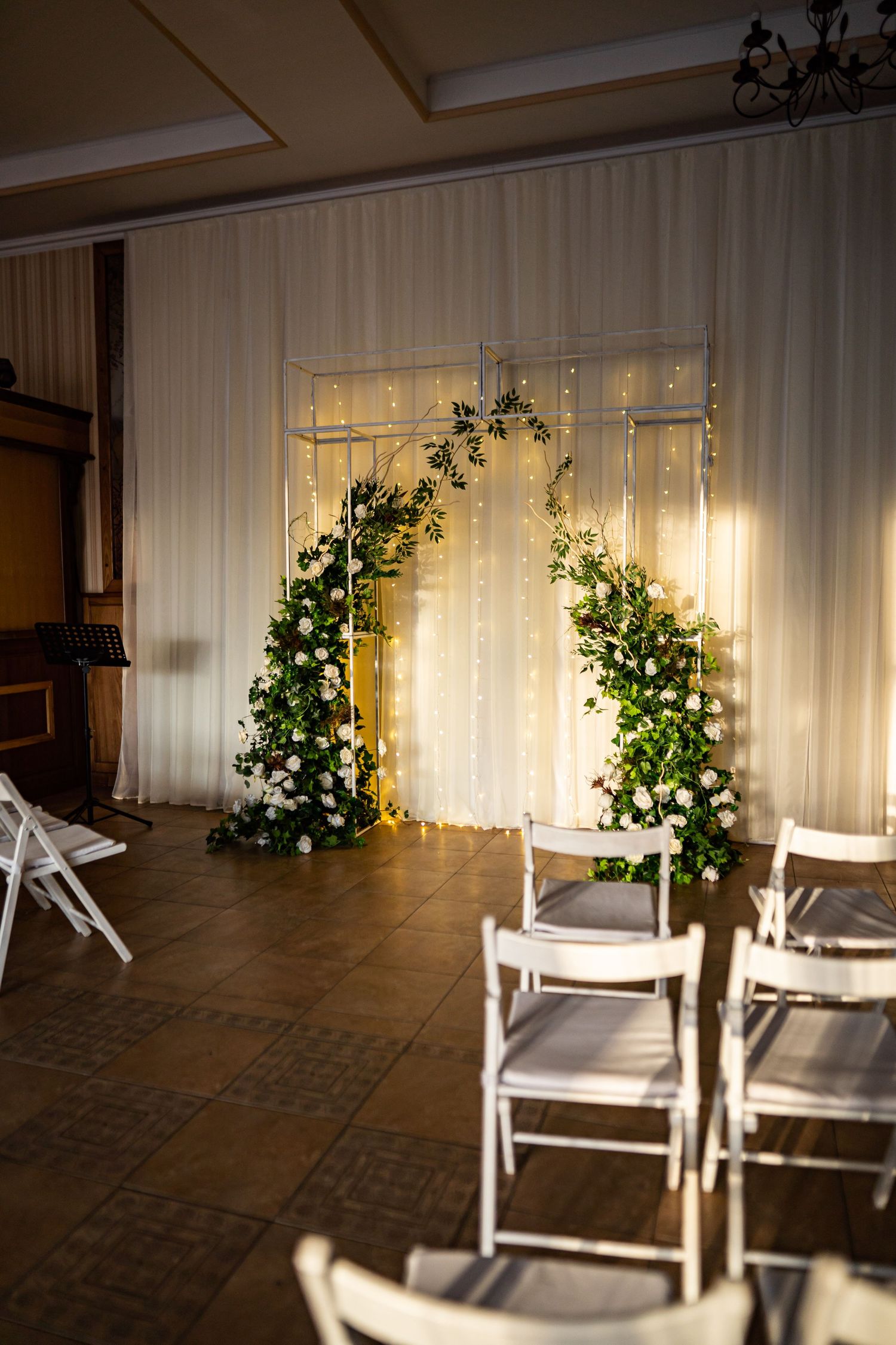 White chairs arranged near sheer curtains with glowing string lights and greenery create an elegant indoor event space.