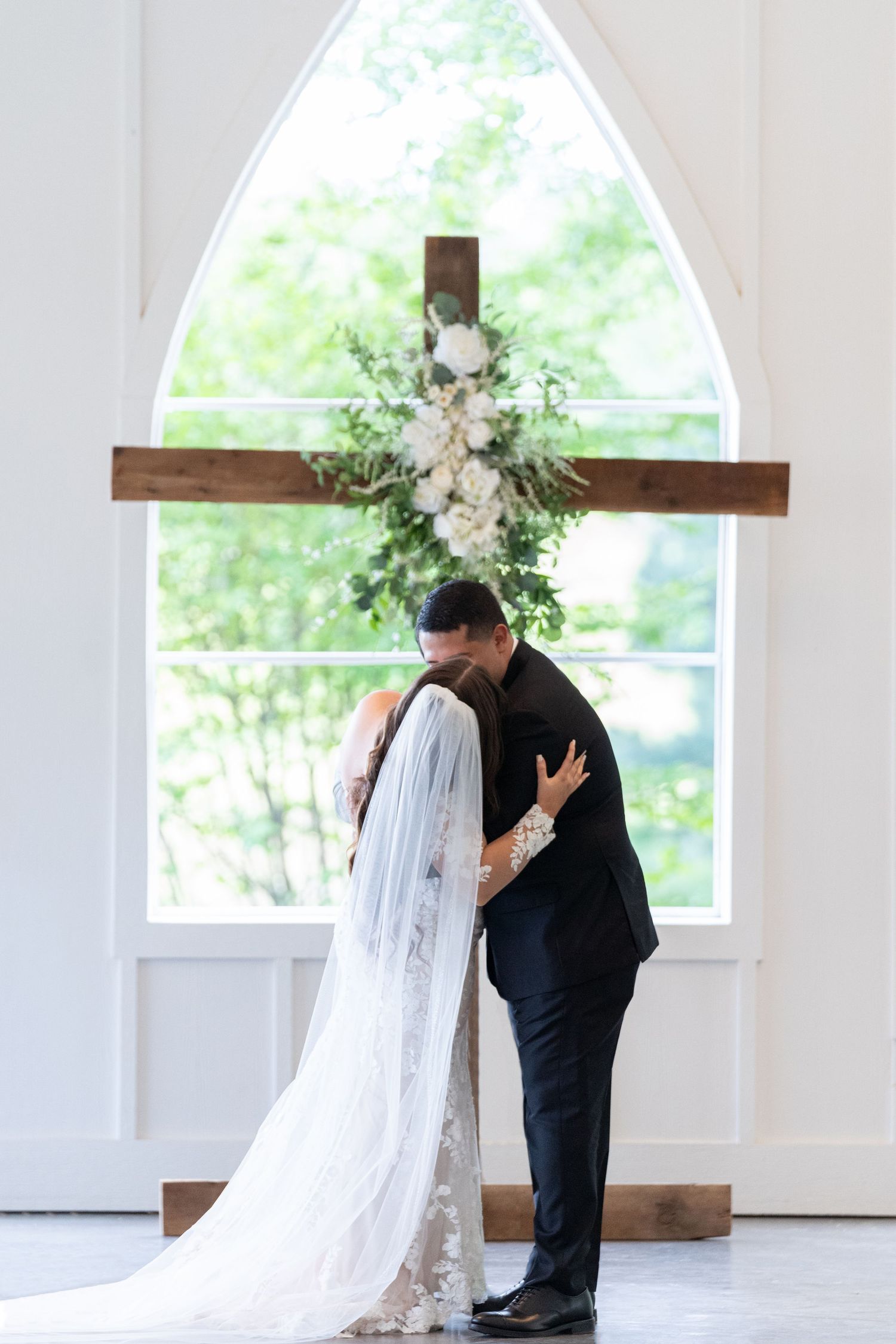 A romantic wedding moment captured in a bright white chapel with arched windows and wooden cross details.