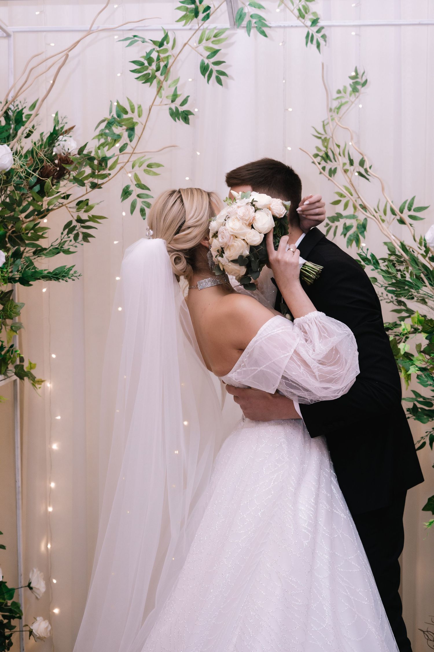 A romantic wedding scene with a couple kissing under a floral arch with twinkling lights and greenery decorations.