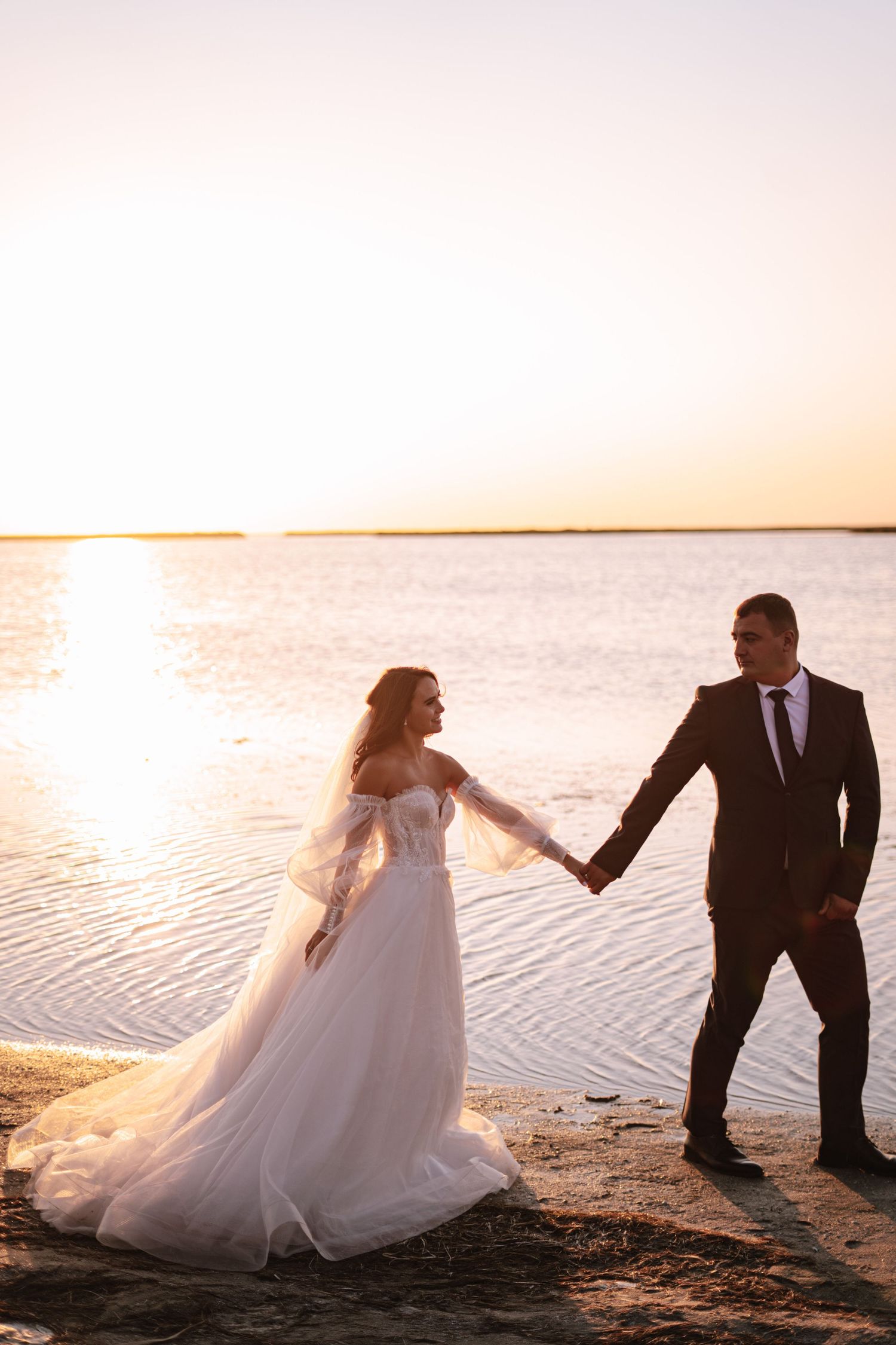 A couple in wedding attire poses playfully on a beach at sunset creating romantic silhouettes against the ocean.