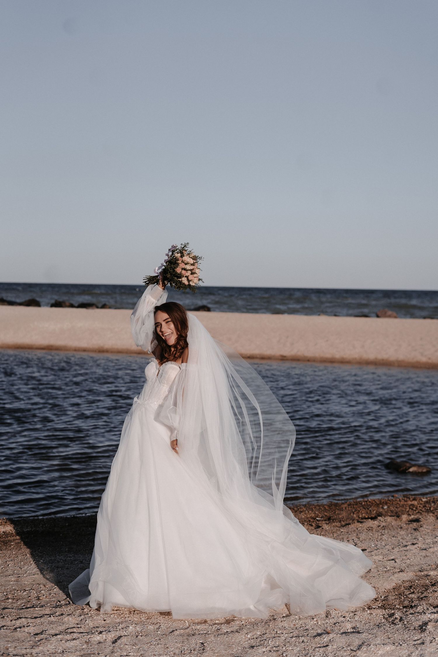 Bride in flowing white wedding dress and long veil holds bouquet while standing on beach at sunset.
