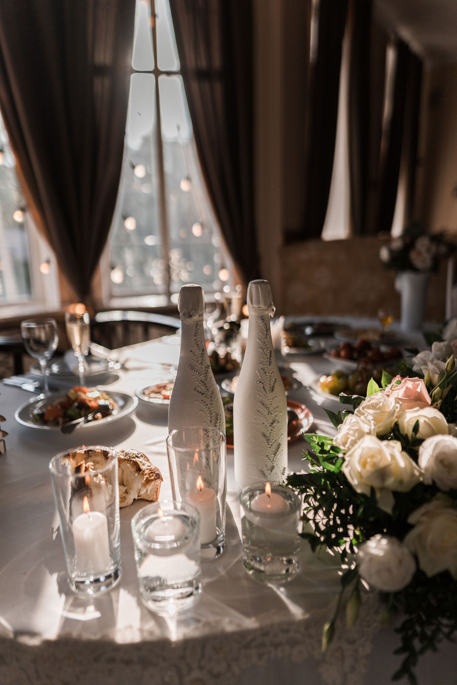 Elegant table setting with frosted glass bottles, candlelight, and white floral arrangements against a window with bokeh lights.