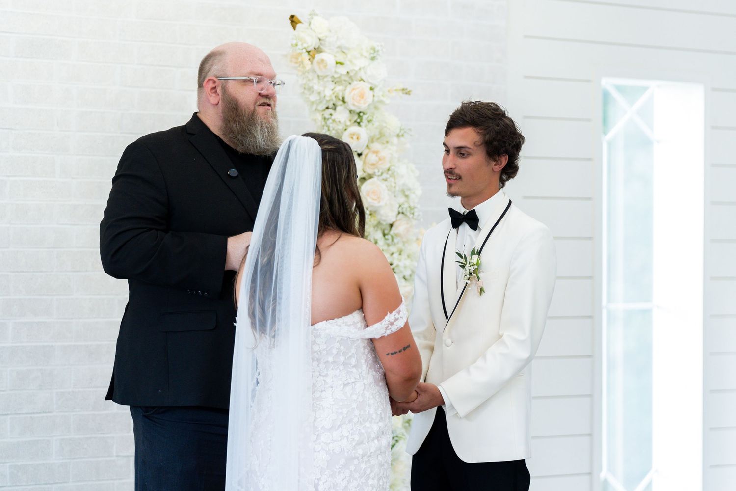 A wedding ceremony takes place in front of a white backdrop with orchids, featuring a bride in white and two people in formal attire.