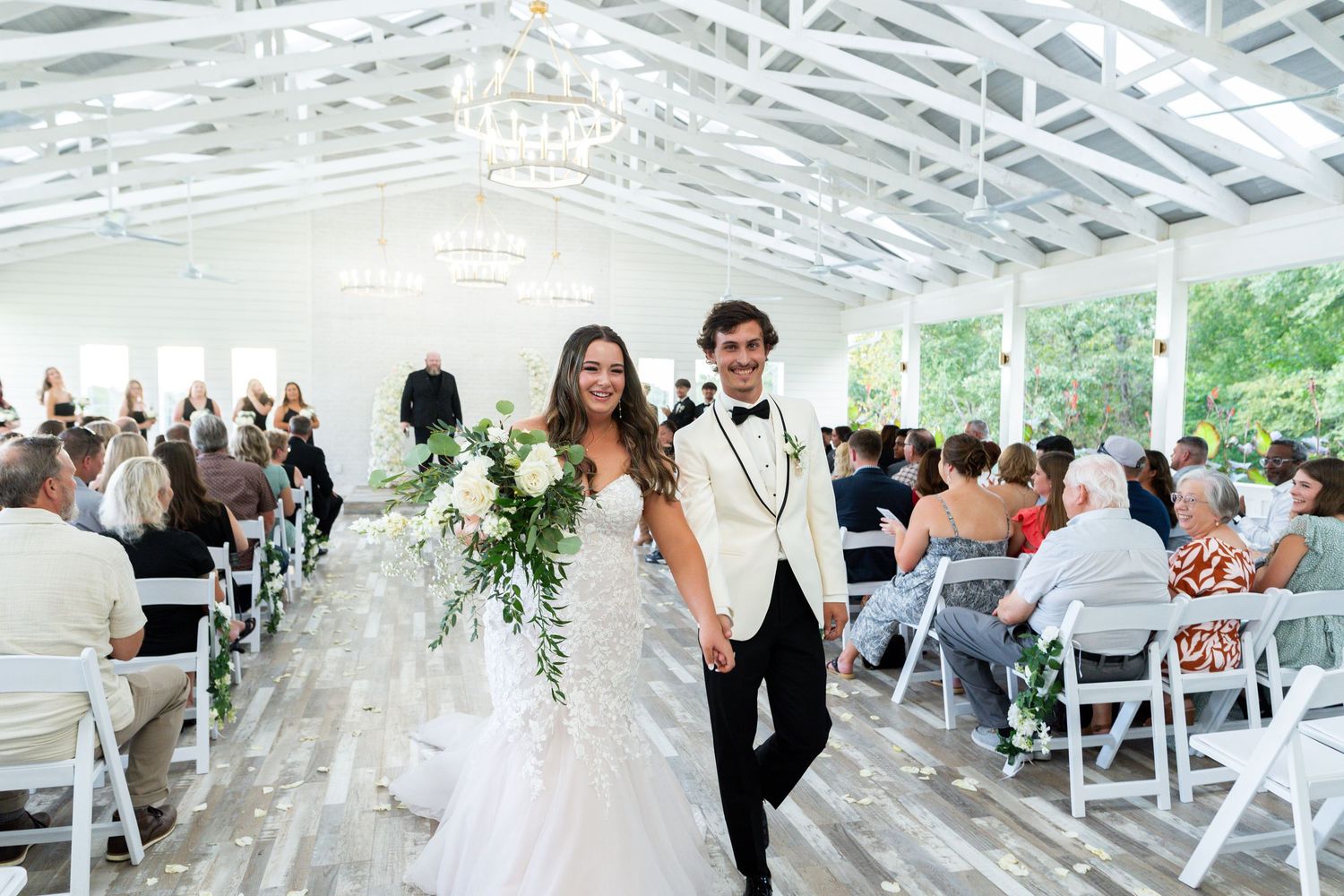 Newlyweds walk down the aisle at a bright greenhouse wedding venue with white chairs and guests in attendance.