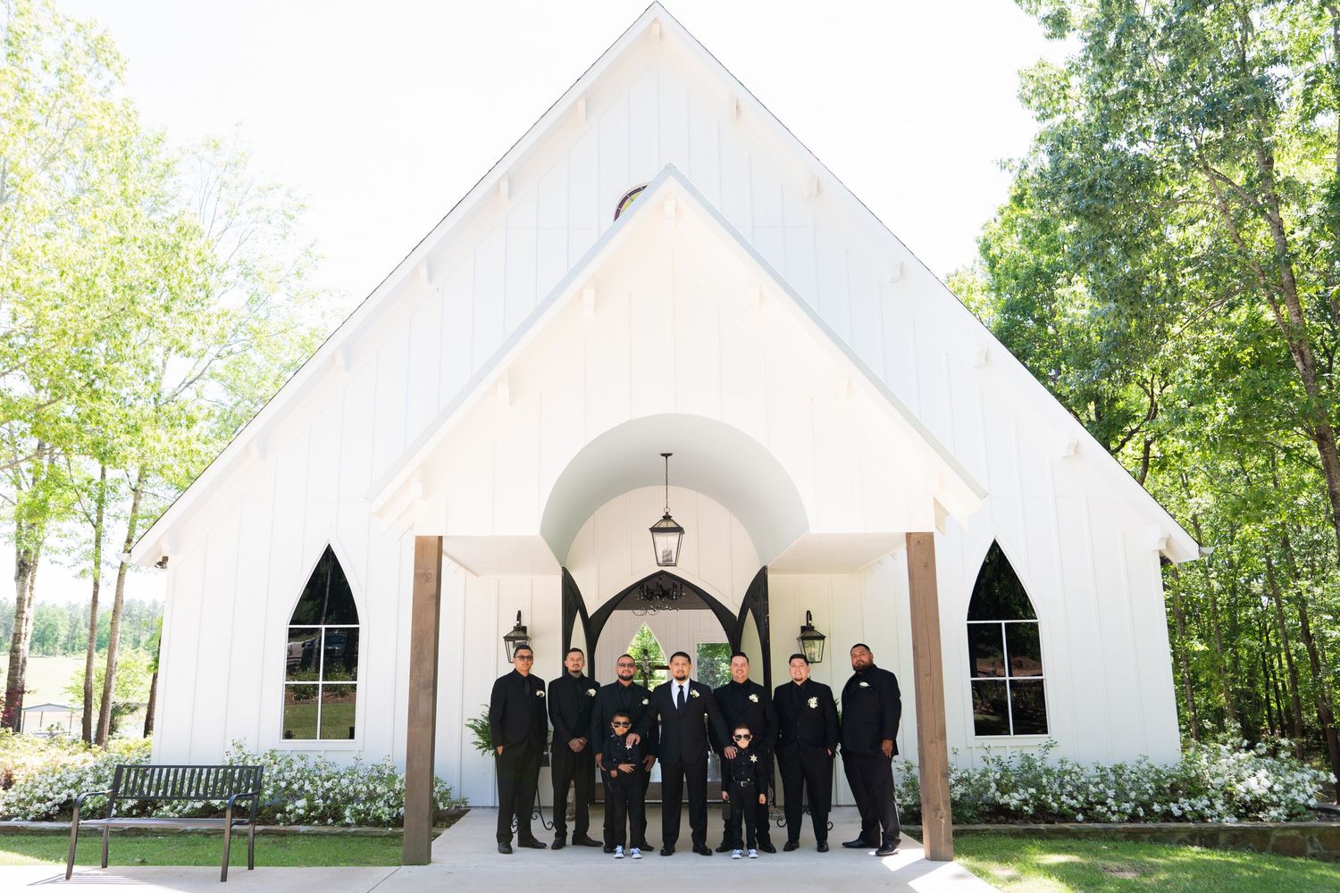 A group of people wearing formal attire stand in front of a white chapel with gothic arched windows surrounded by greenery.
