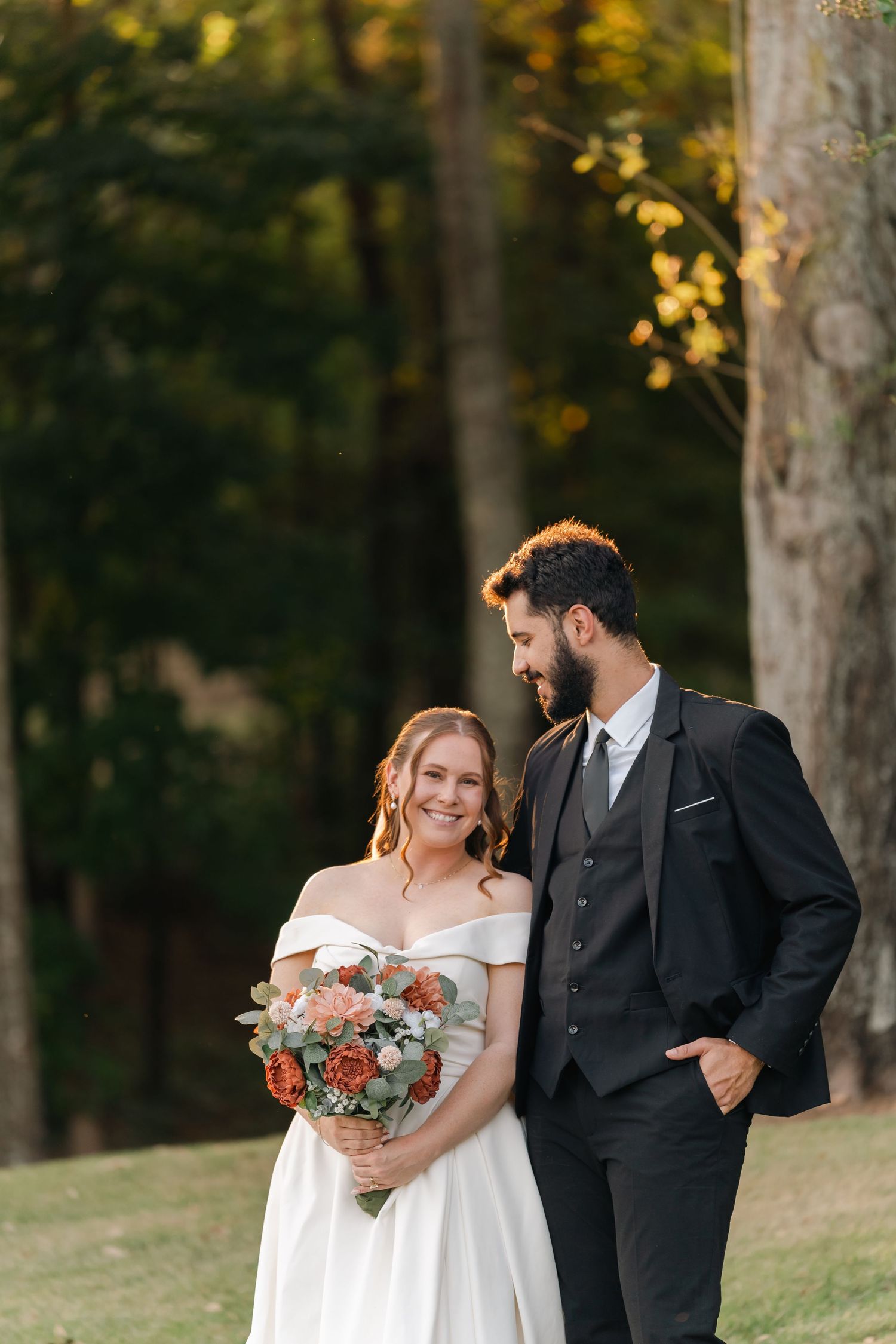 A romantic outdoor wedding portrait shows a couple standing among trees at sunset with the bride holding a floral bouquet.