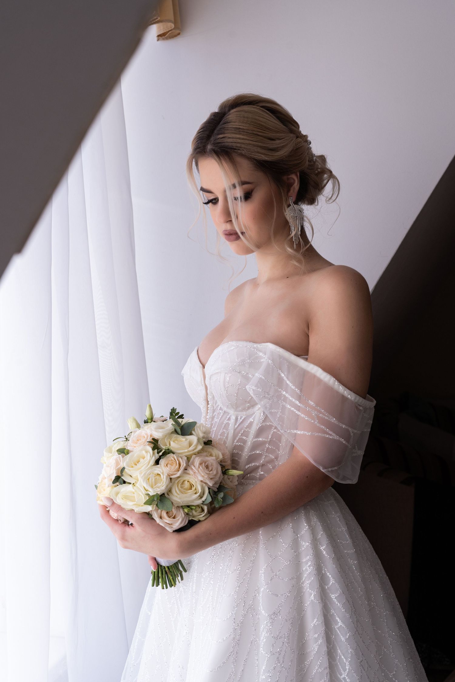 A bride in a flowing white wedding gown holds a bouquet of cream roses against a light backdrop.