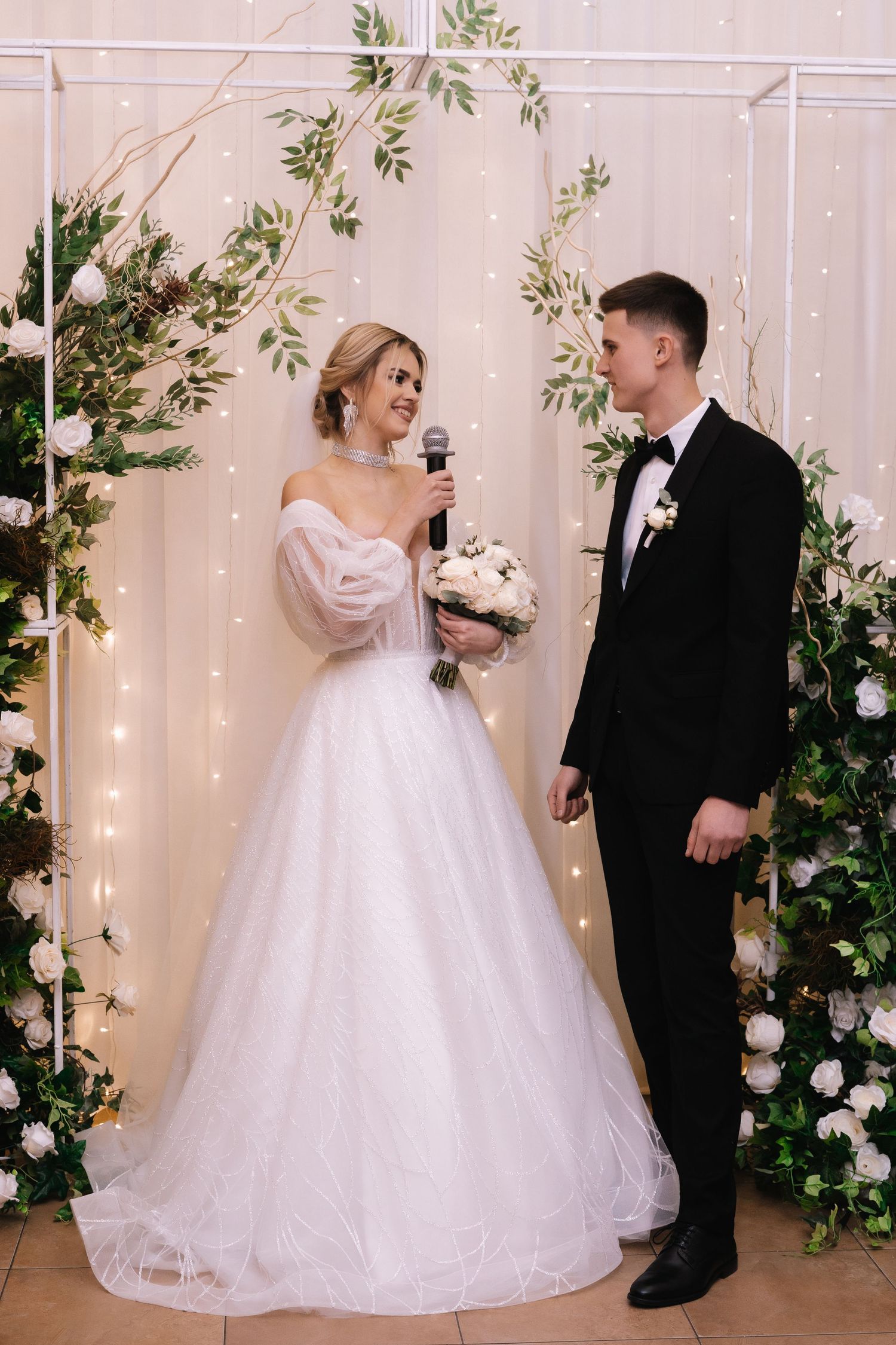 Bride and groom share a romantic moment in front of a floral backdrop with twinkling lights during their wedding ceremony.