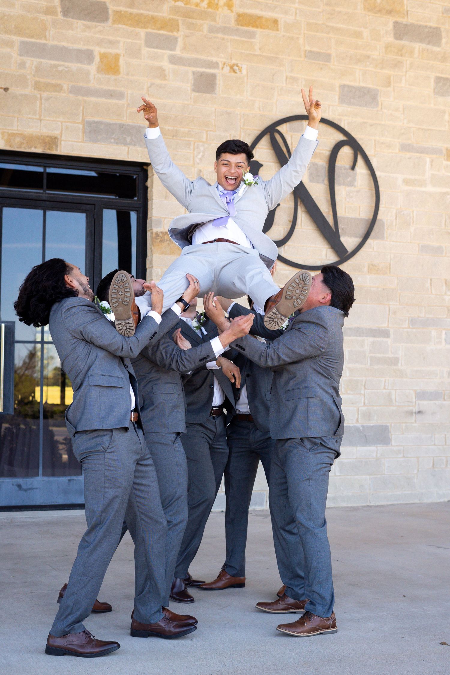 Groomsmen in grey suits playfully posing and jumping outside a brick building.