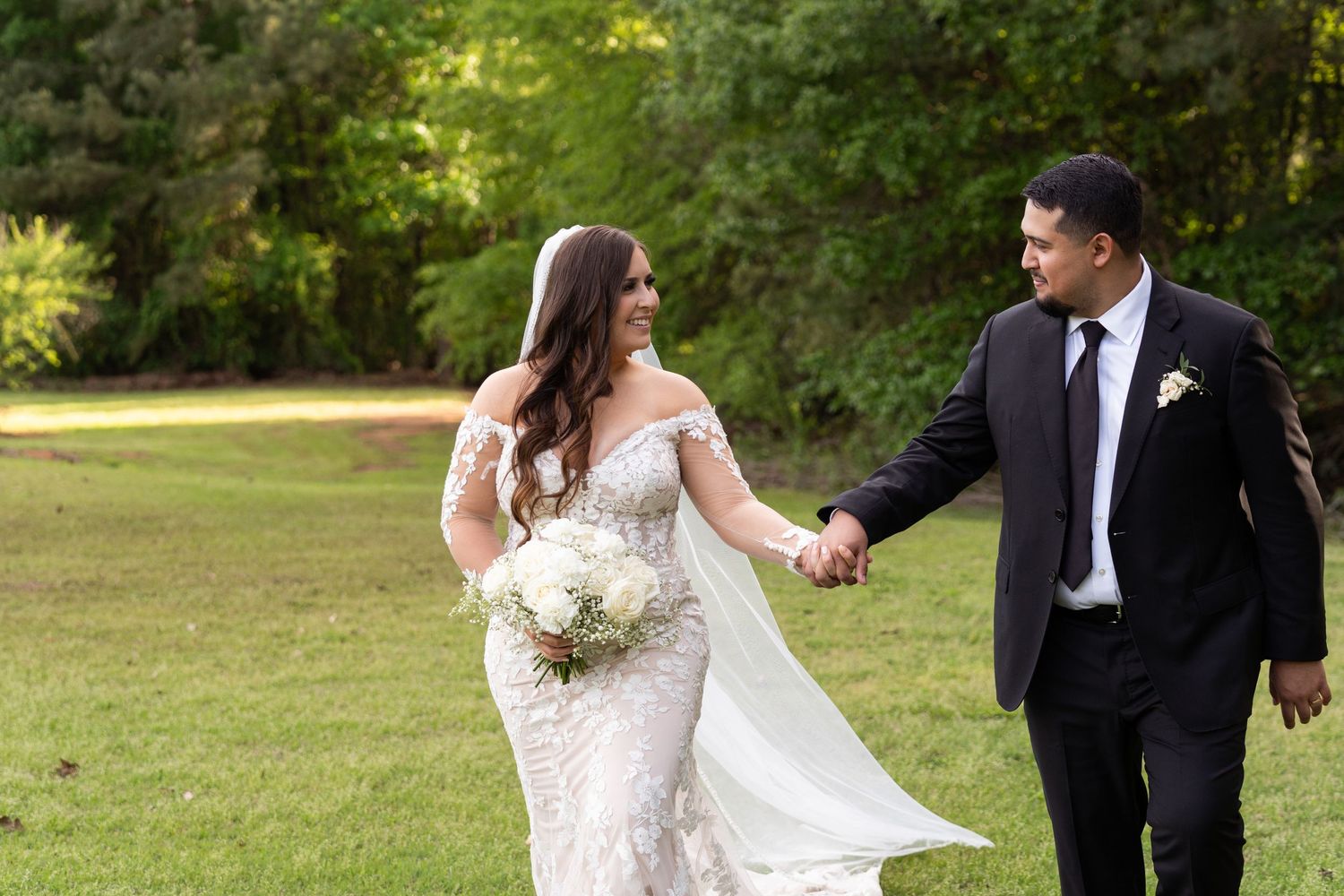 A couple holds hands while walking on a grassy lawn, with the bride wearing a fitted lace gown and holding a bouquet.