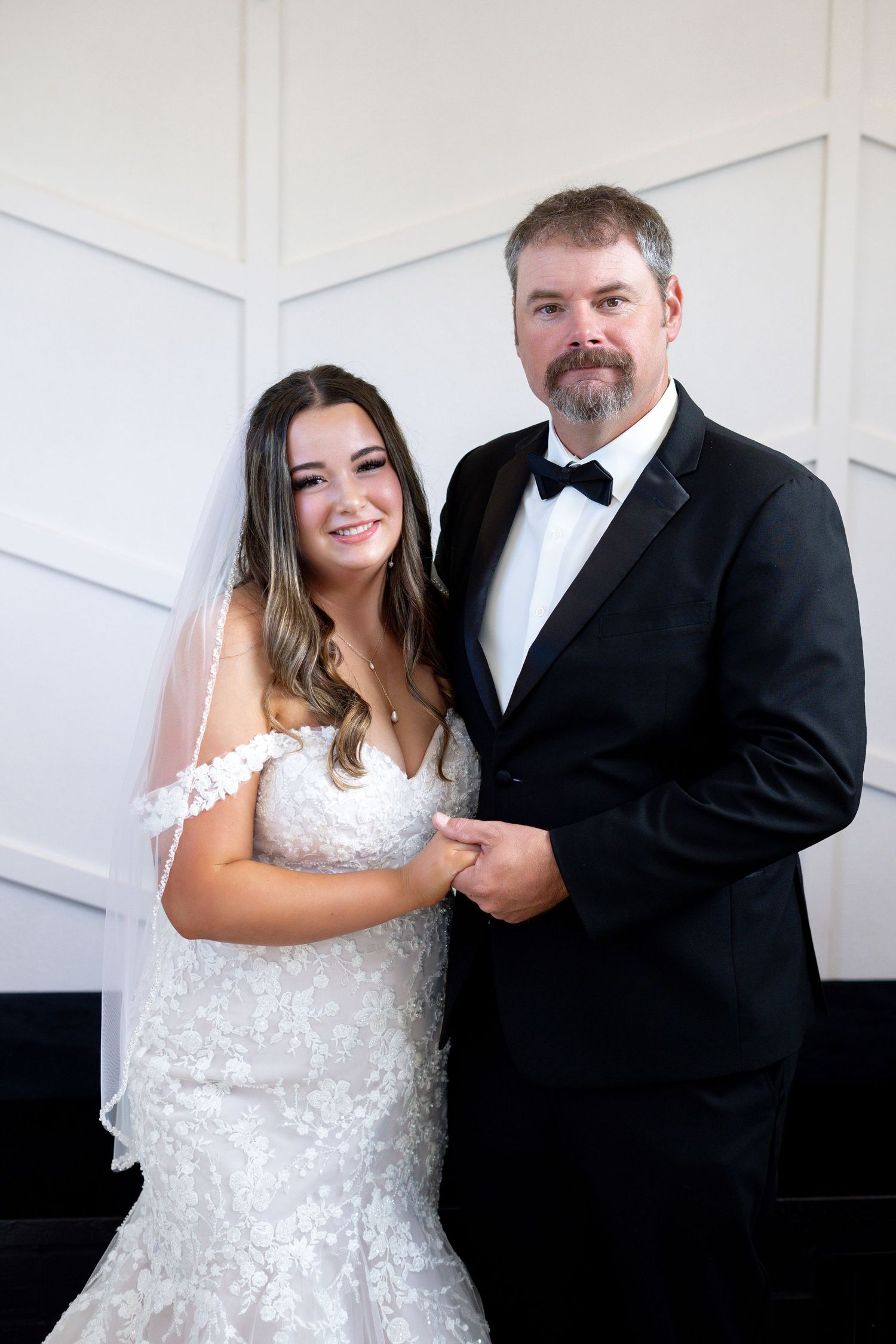 A bride in a white lace wedding dress and groom in black tuxedo pose together against a white paneled wall.
