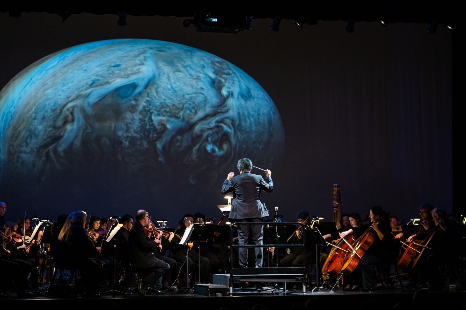 Symphony orchestra performs beneath large celestial projections in a darkened concert hall.