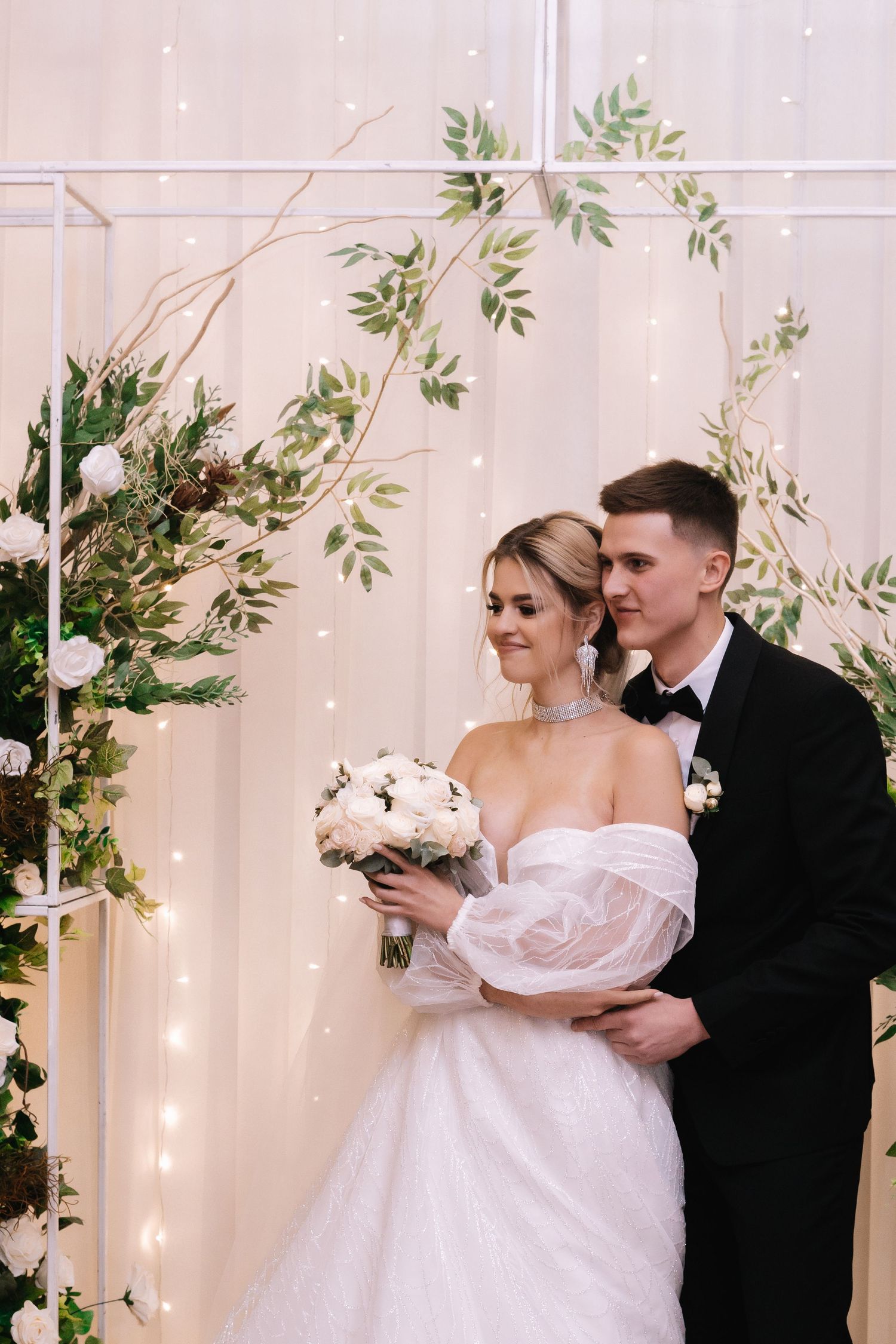 Elegant wedding couple stands against light backdrop with greenery and twinkle lights while holding white floral bouquet.