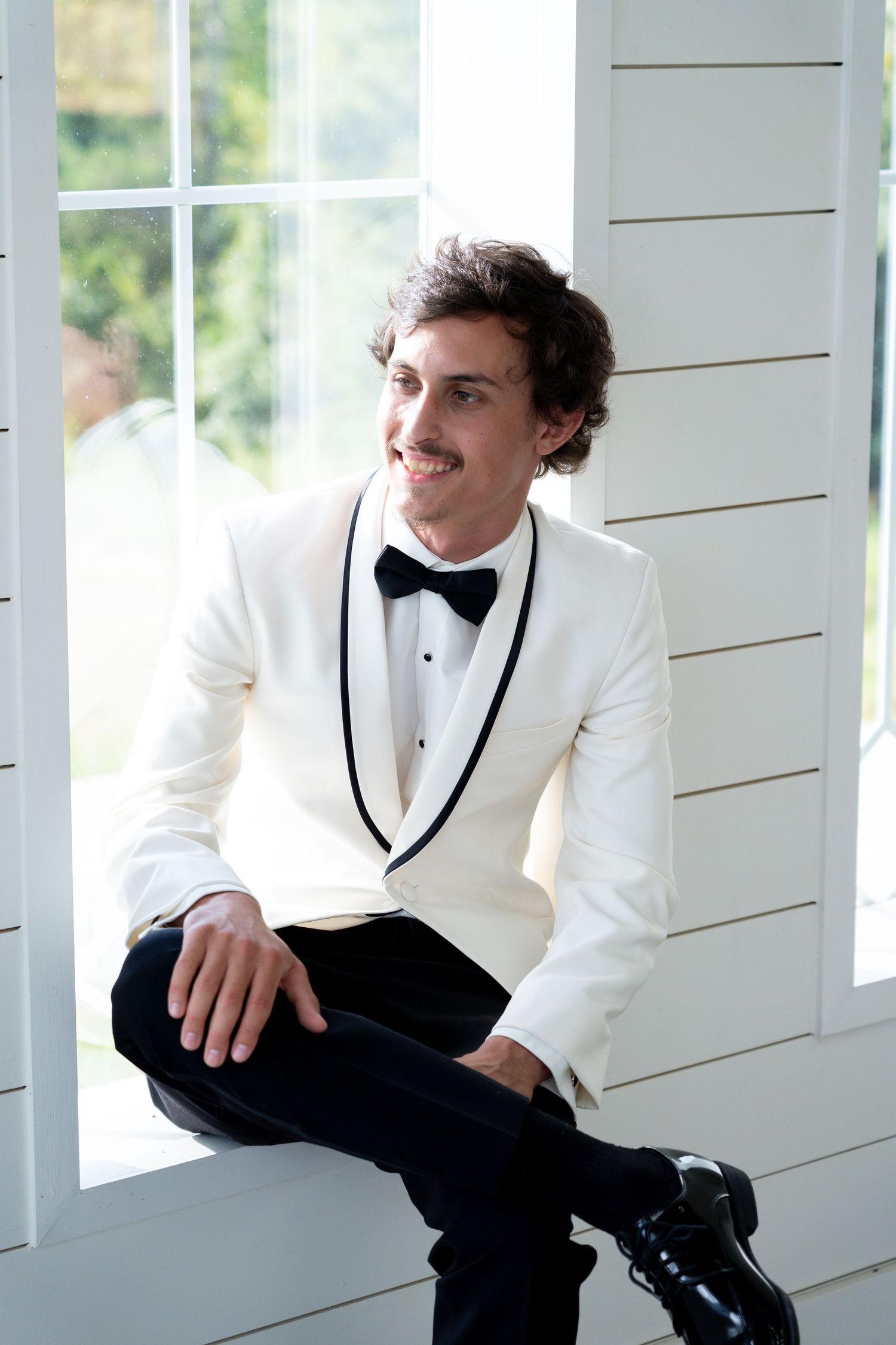 A person in a white tuxedo jacket and black bowtie sits casually on a white porch railing in an elegant setting.
