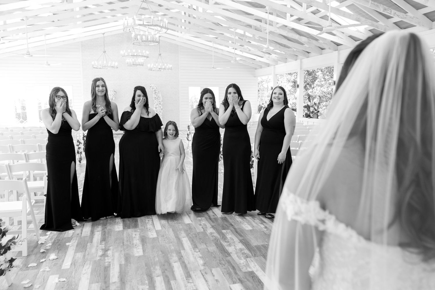 A bridal party in black bridesmaid dresses reacts with joy as they see the bride in her white gown and veil.