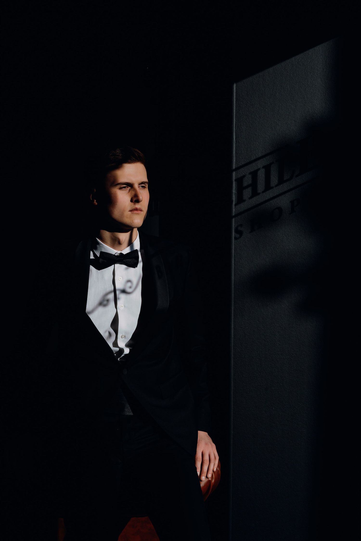 A dramatic portrait in dark lighting shows someone in formal attire with a bow tie standing against a shadowy wall.