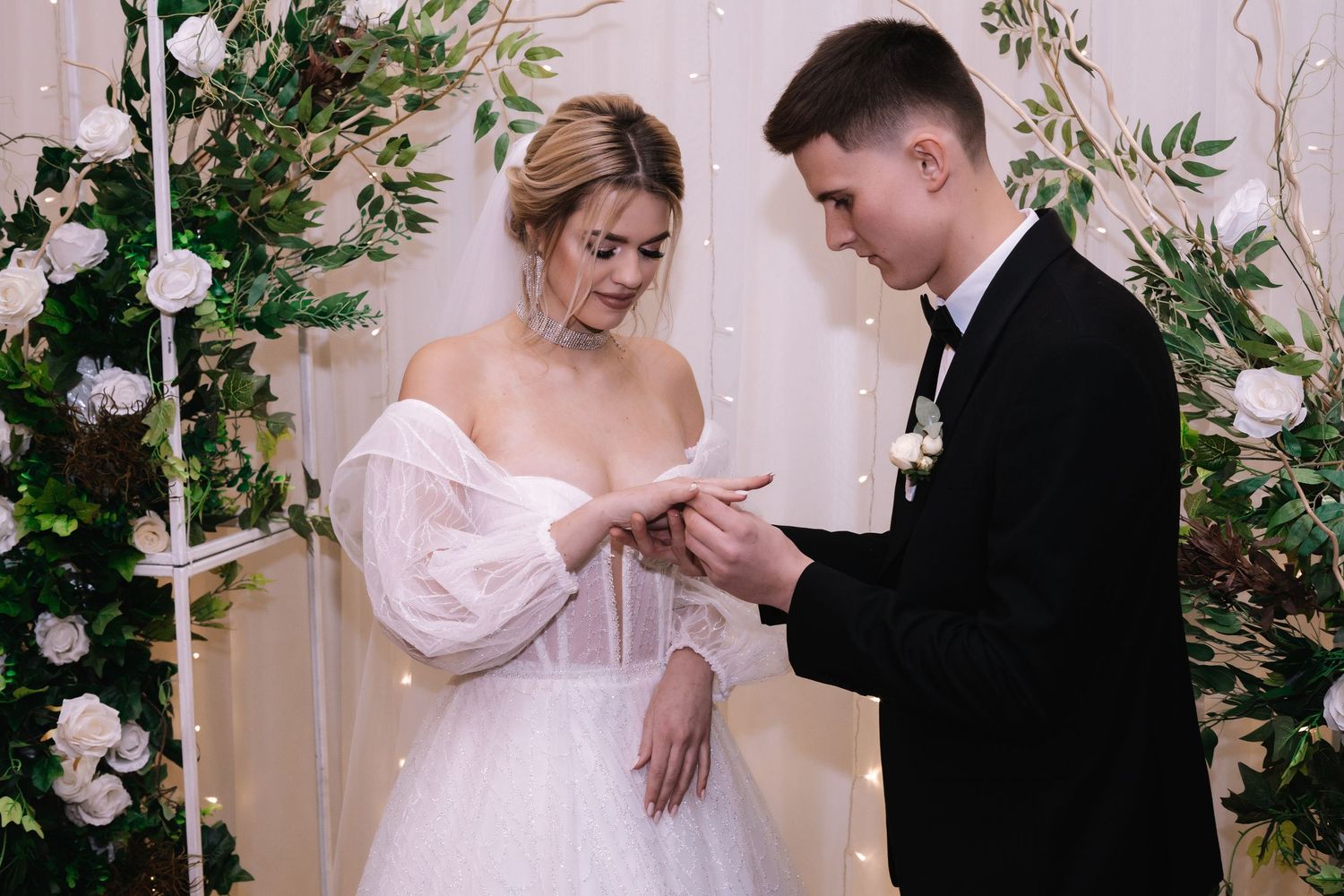 A romantic wedding ceremony moment with white floral decor and greenery as two people exchange rings.