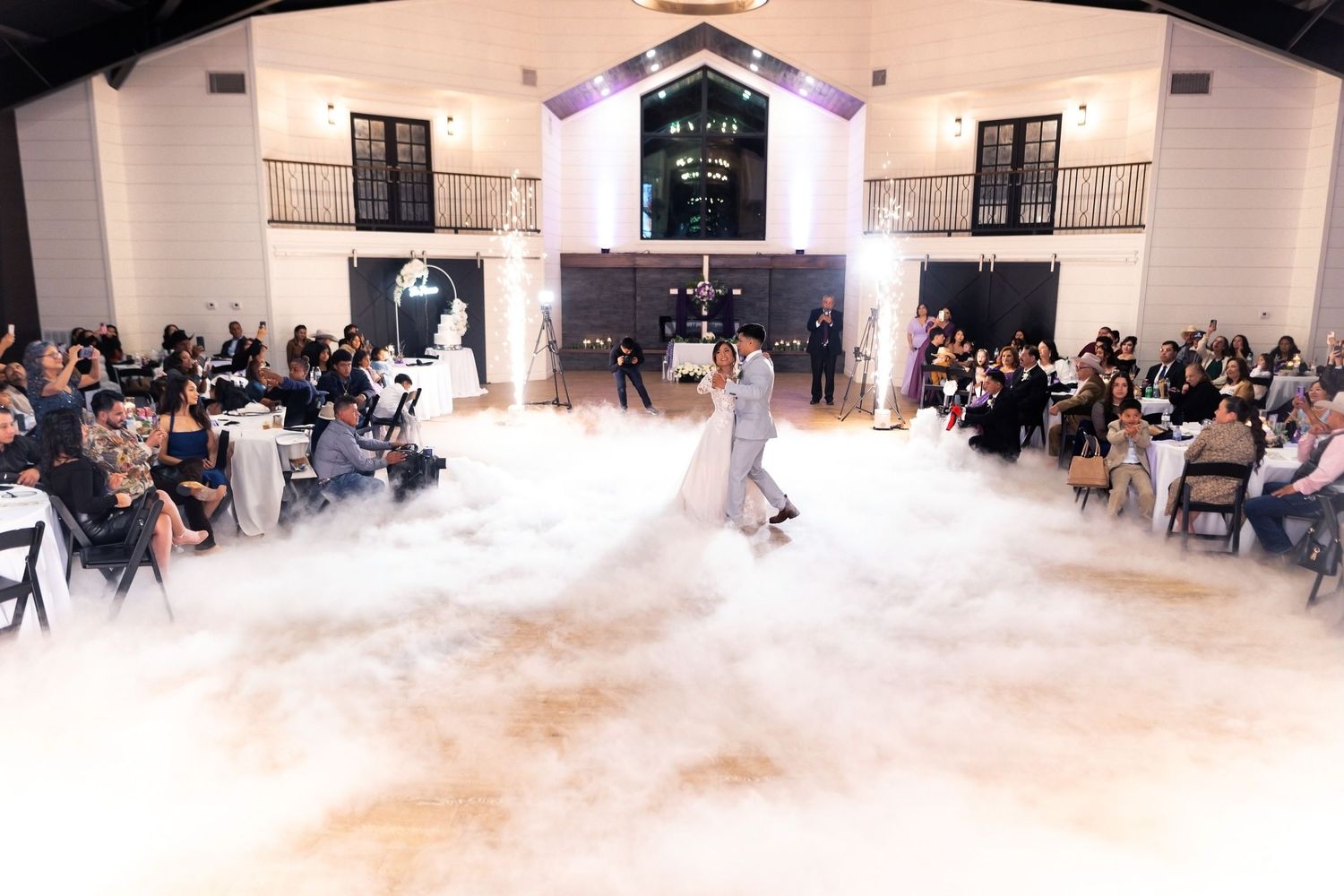 A large crowd gathers in a white event space as fog machines create a dramatic low-lying mist effect across the floor.