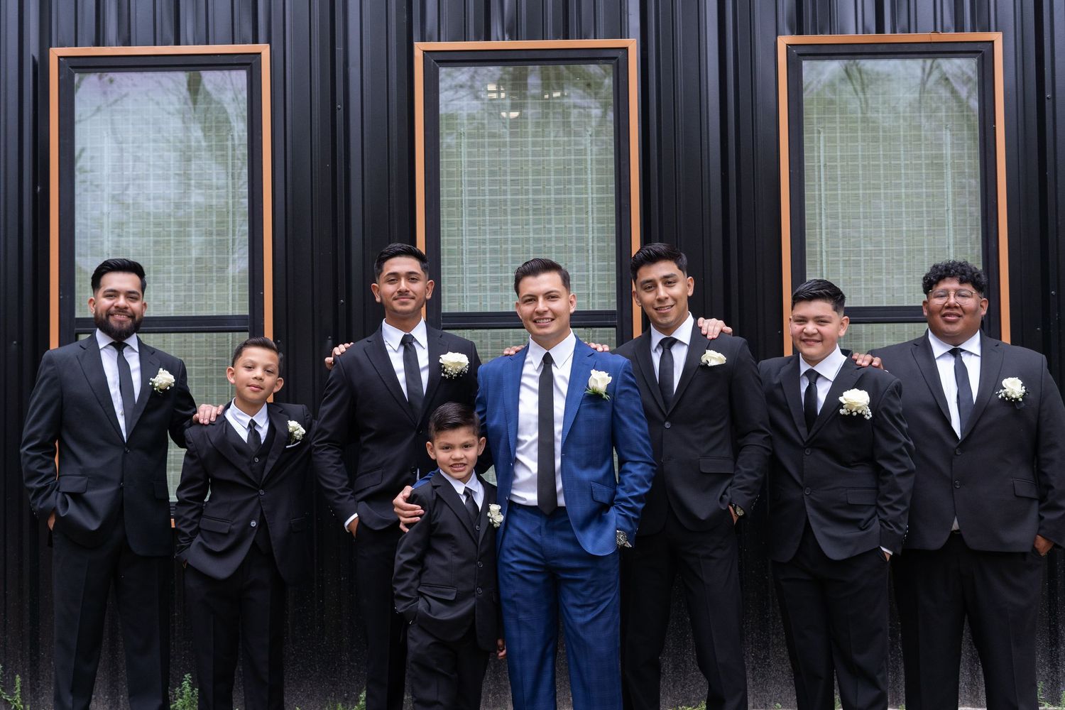 A wedding party in formal attire stands in a line against a dark striped wall with windows.