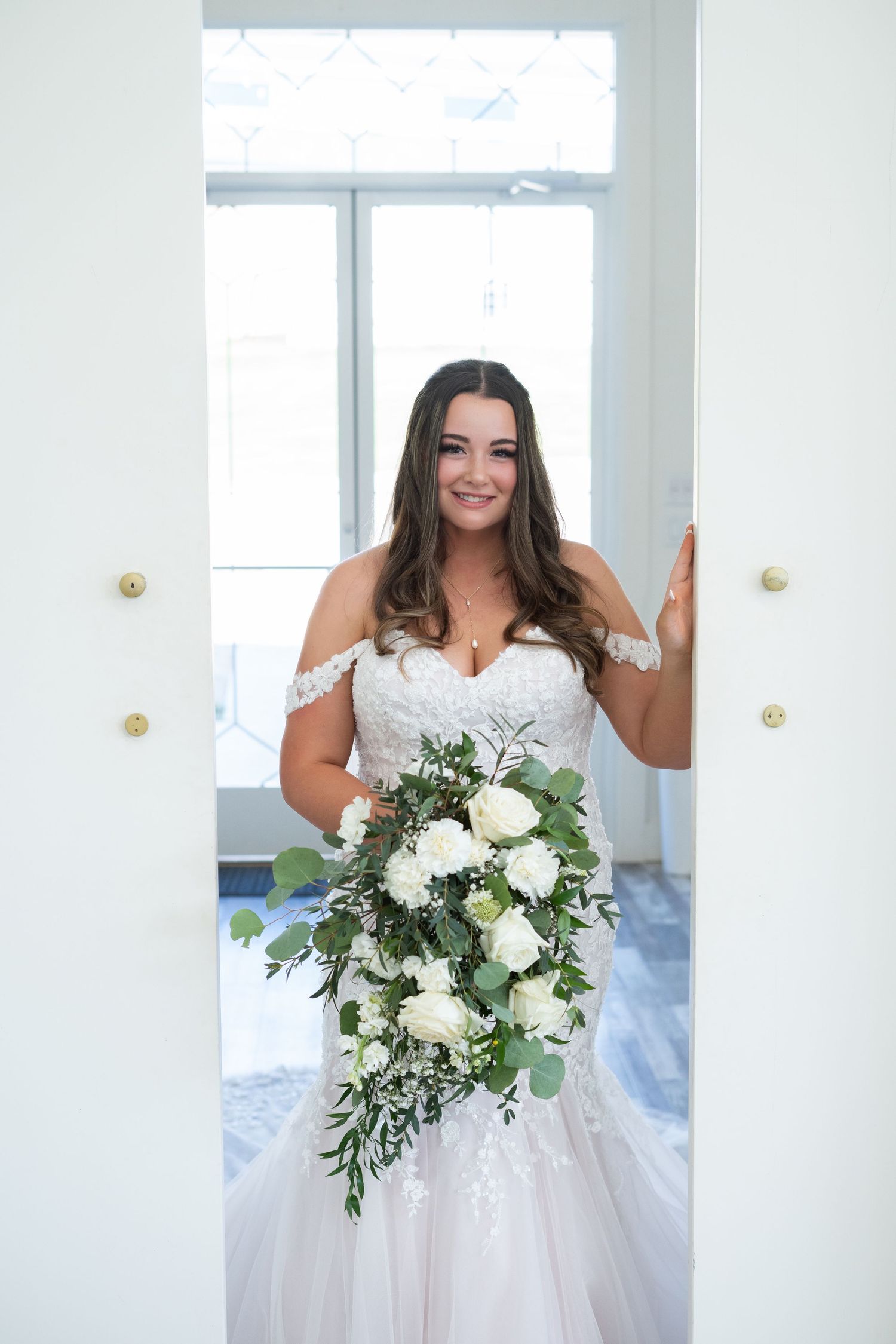 Beautiful bridal portrait with cascading white floral bouquet and off-shoulder lace wedding gown in bright indoor setting.