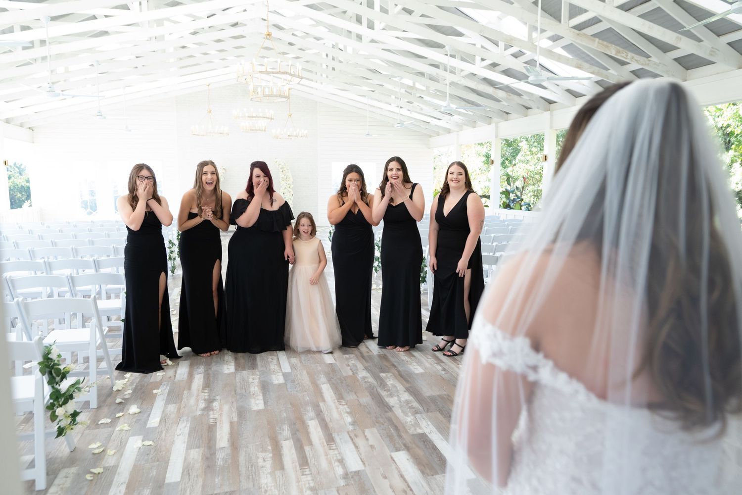 A wedding party in black bridesmaid dresses stands in a bright covered venue with a veiled figure in the foreground.