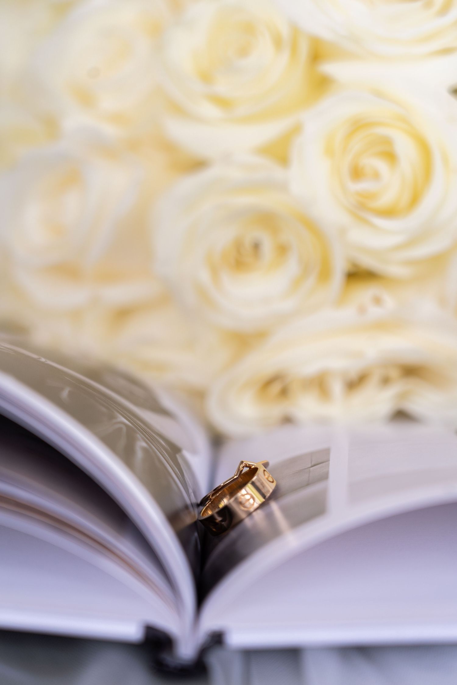 A gold wedding ring rests between pages of an open book with soft white roses in the background.