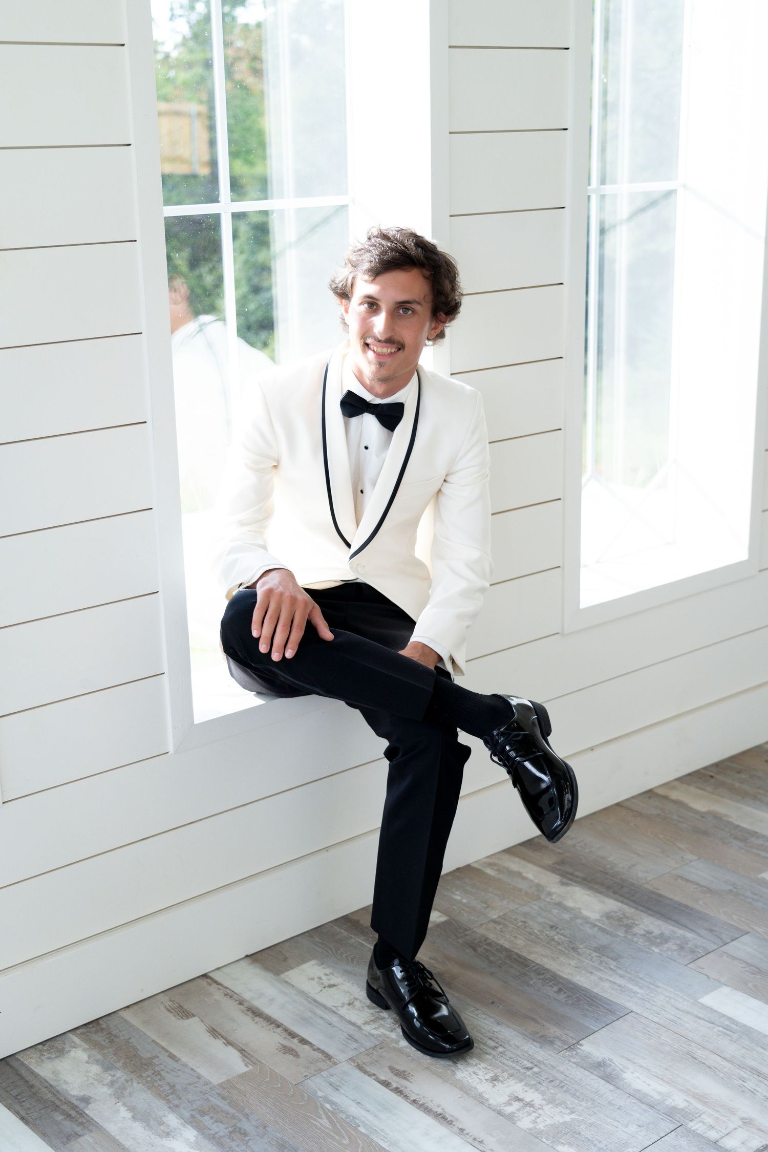 A person in formal black and white attire poses in a bright white room with wooden floors.