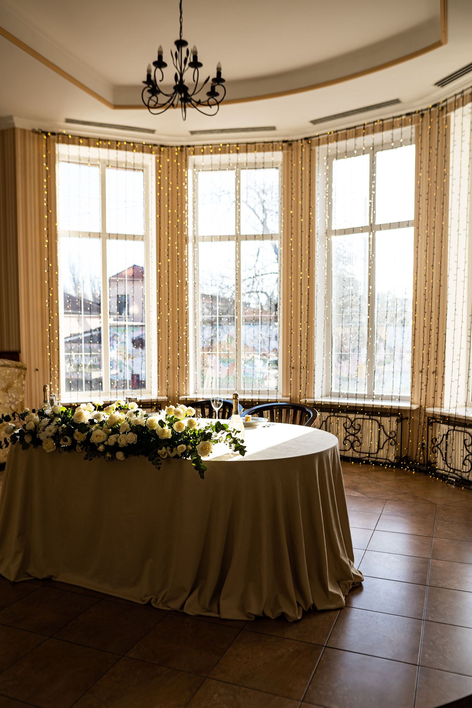 Elegant banquet room with bay windows, chandelier, and long draped table decorated with floral arrangements and soft lighting.